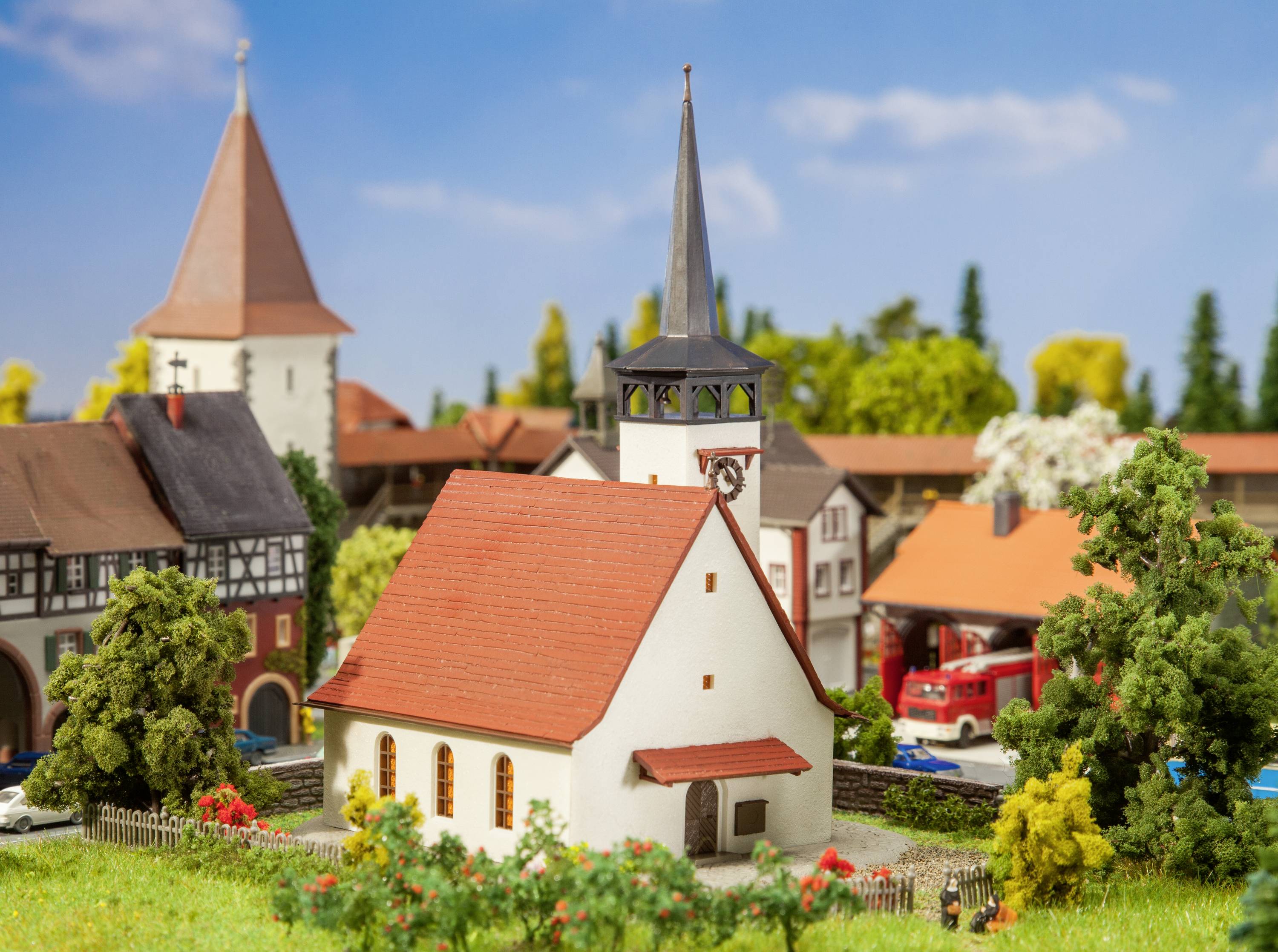 Model van een schilderachtige dorpskerk met een rood dak naast een brandweerwagen in een miniatuurdorp met meerdere gebouwen en bomen.