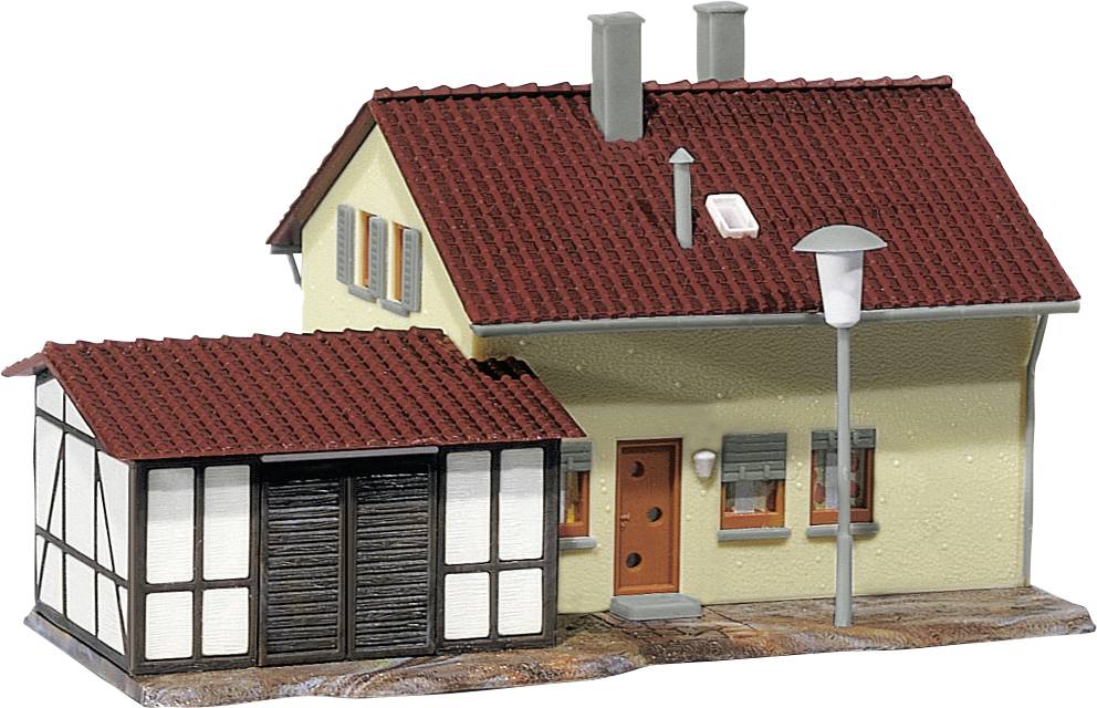 Een modelhuis met een rood dak en een geel-bruine gevel, bijgebouwen met houten bekleding, en een straatlantaarn ervoor.