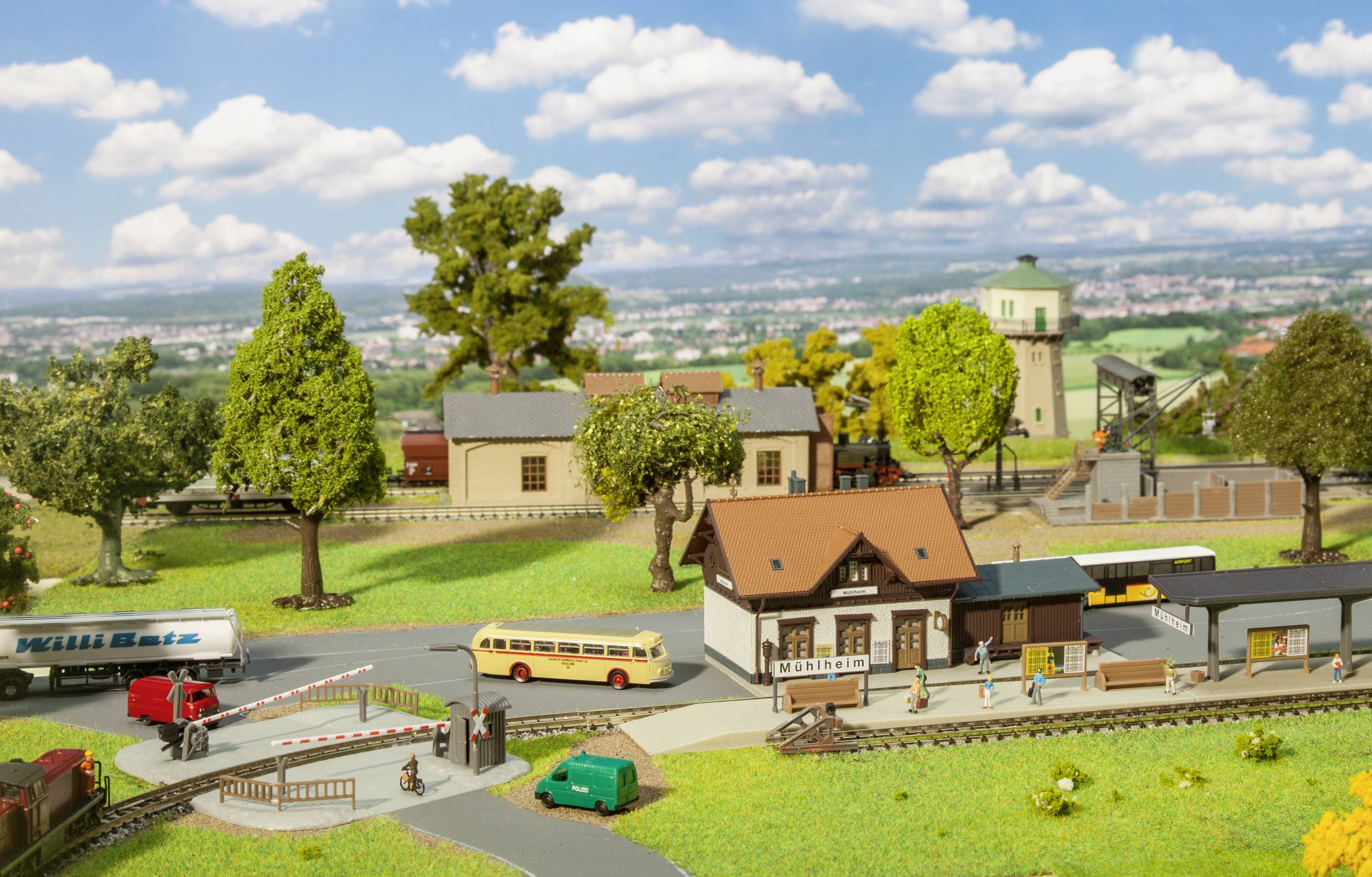 Klein model van een plattelandsstation met trein, bus, vrachtwagen en gebouwen, omgeven door bomen en landschap op de achtergrond.