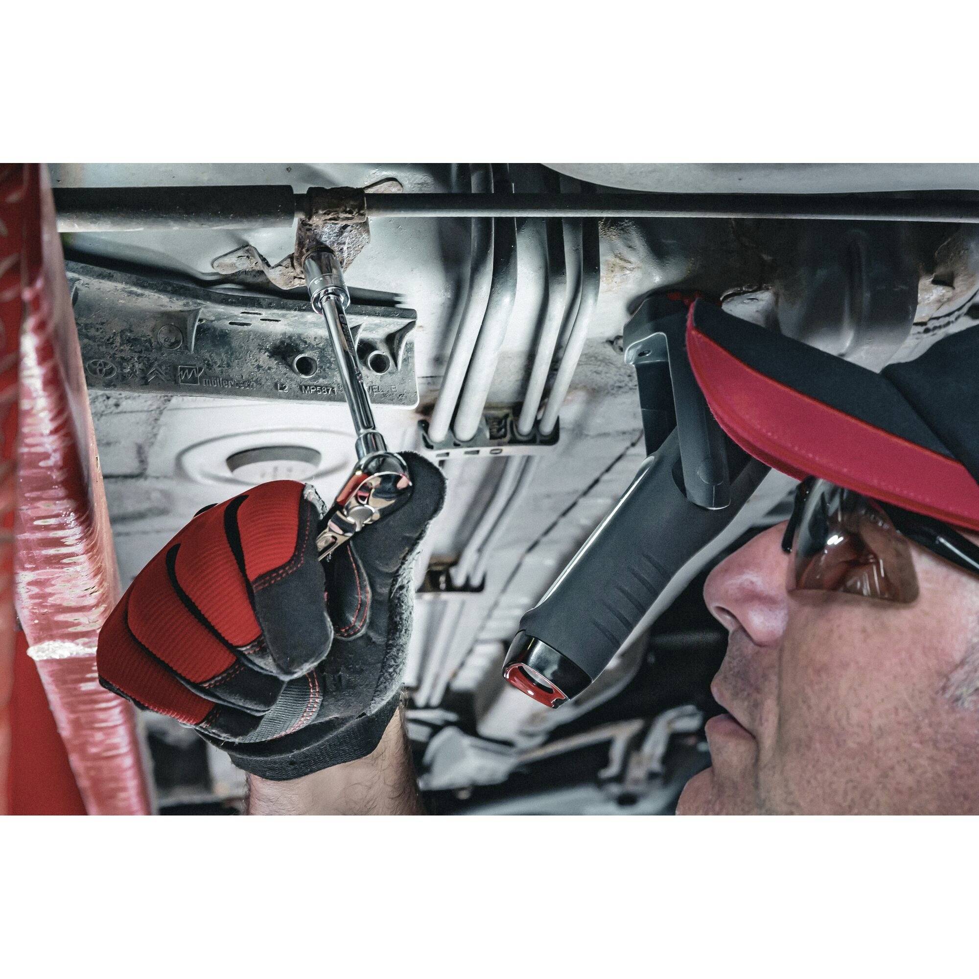 Een monteur in rode handschoen en veiligheidsbril repareert een auto met een moersleutel onder een voertuig.