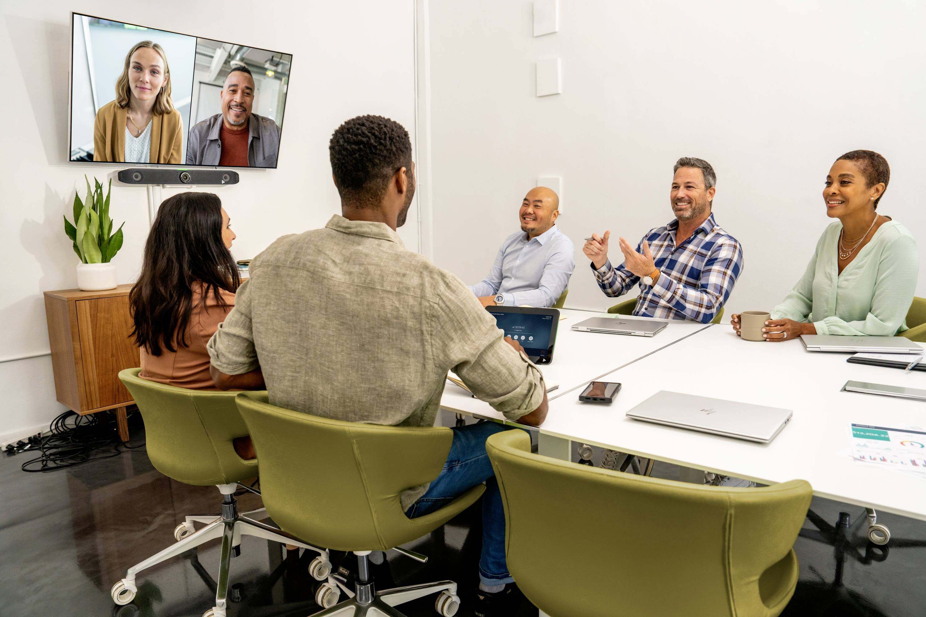 Zes personen in een vergaderruimte, vier aan tafel, twee op een scherm via videoconferentie, discussiëren lachend.