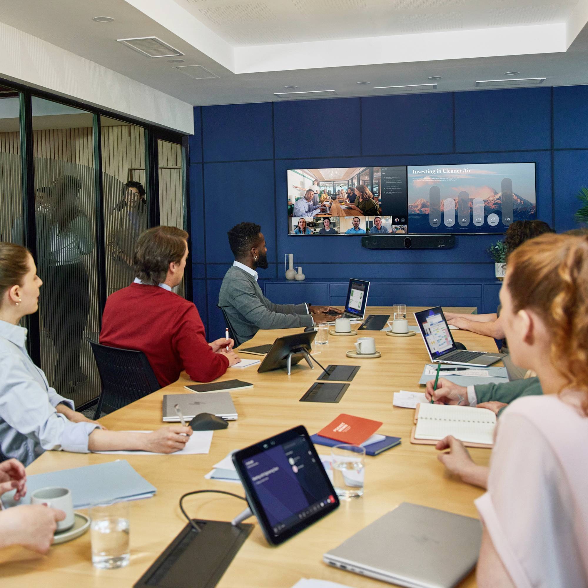 Mensen zitten in een vergaderkamer met laptops en aantekeningen terwijl ze deelnemen aan een videoconferentie op een scherm.