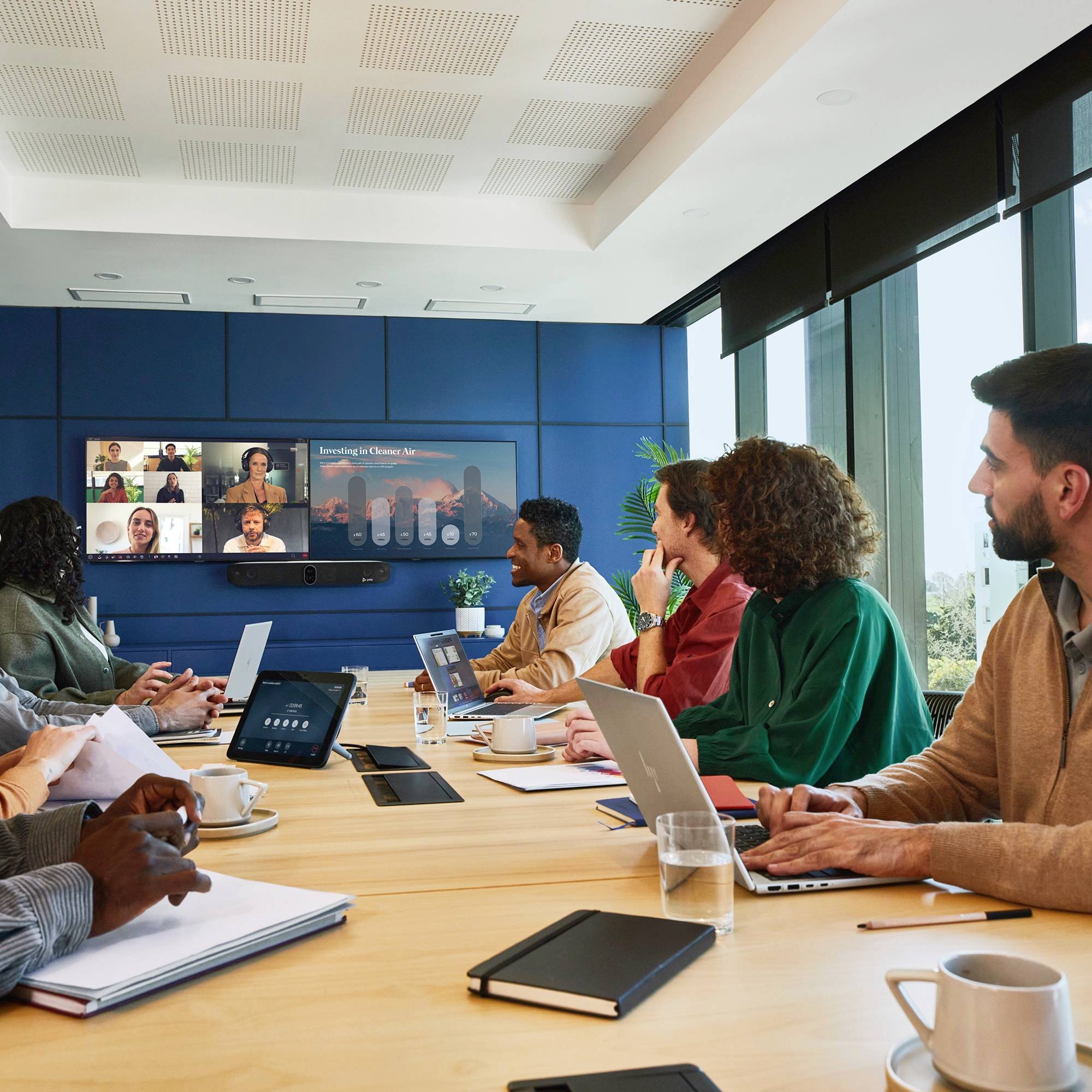 Mensen in een vergaderruimte nemen deel aan een hybride vergadering. Enkele deelnemers zijn zichtbaar op een videoconferentie-scherm.