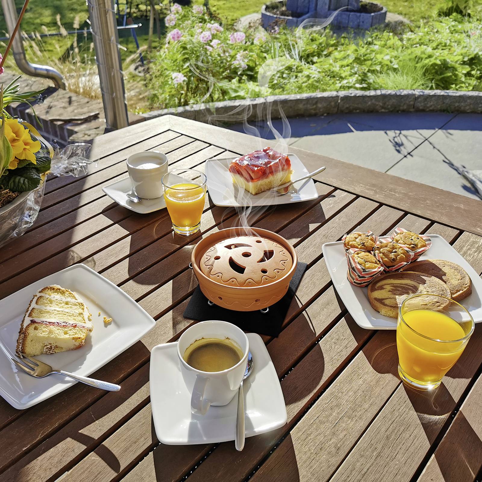 Ontbijttafel buiten met cake, koekjes, koffie en sinaasappelsap. In het midden een rokende terracotta pot als theelichthouder. Tuin op de achtergrond.