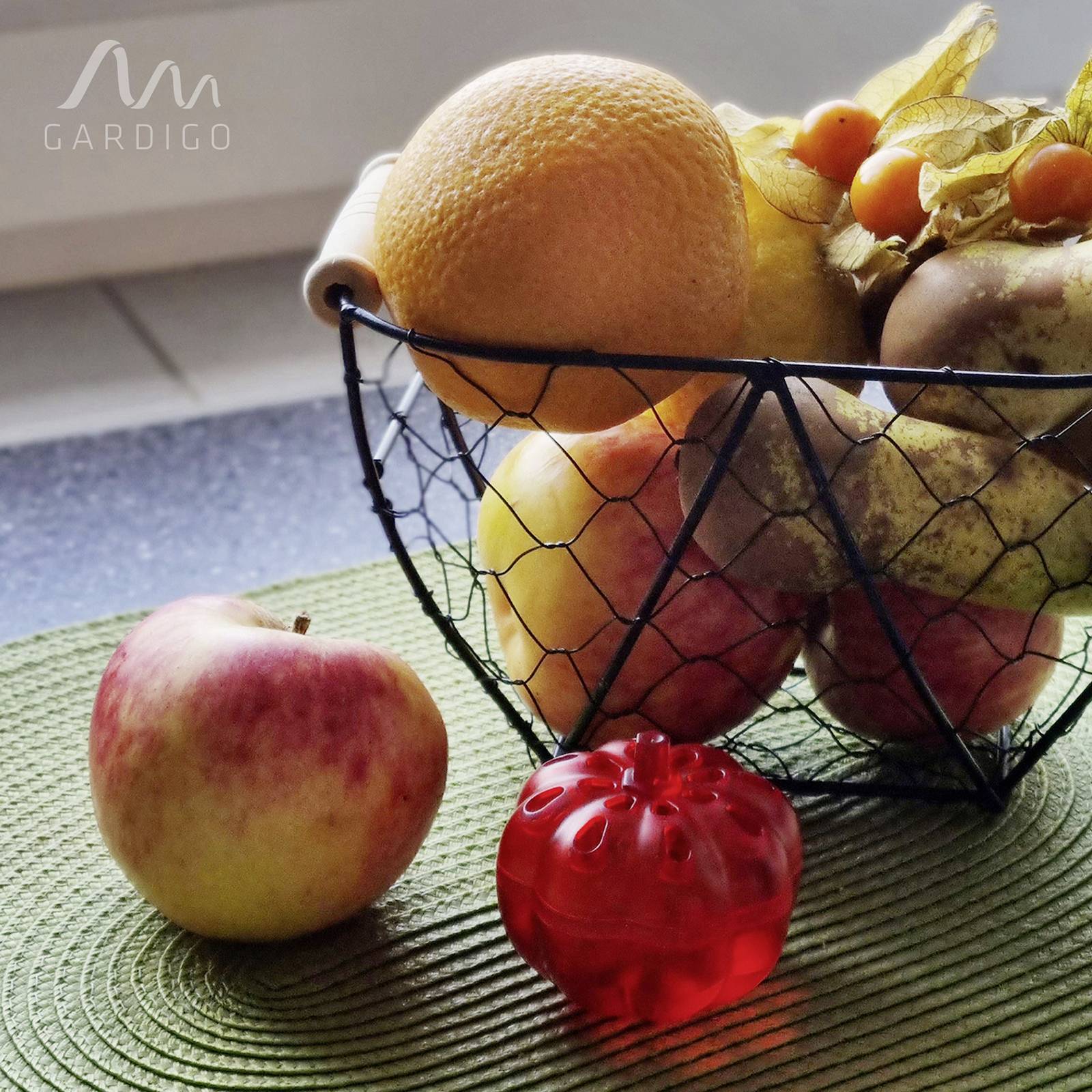 Een draadmand met lampionplanten, peren en sinaasappels. Op de voorgrond een appel en een rode decoratieve figuur op een groene mat.
