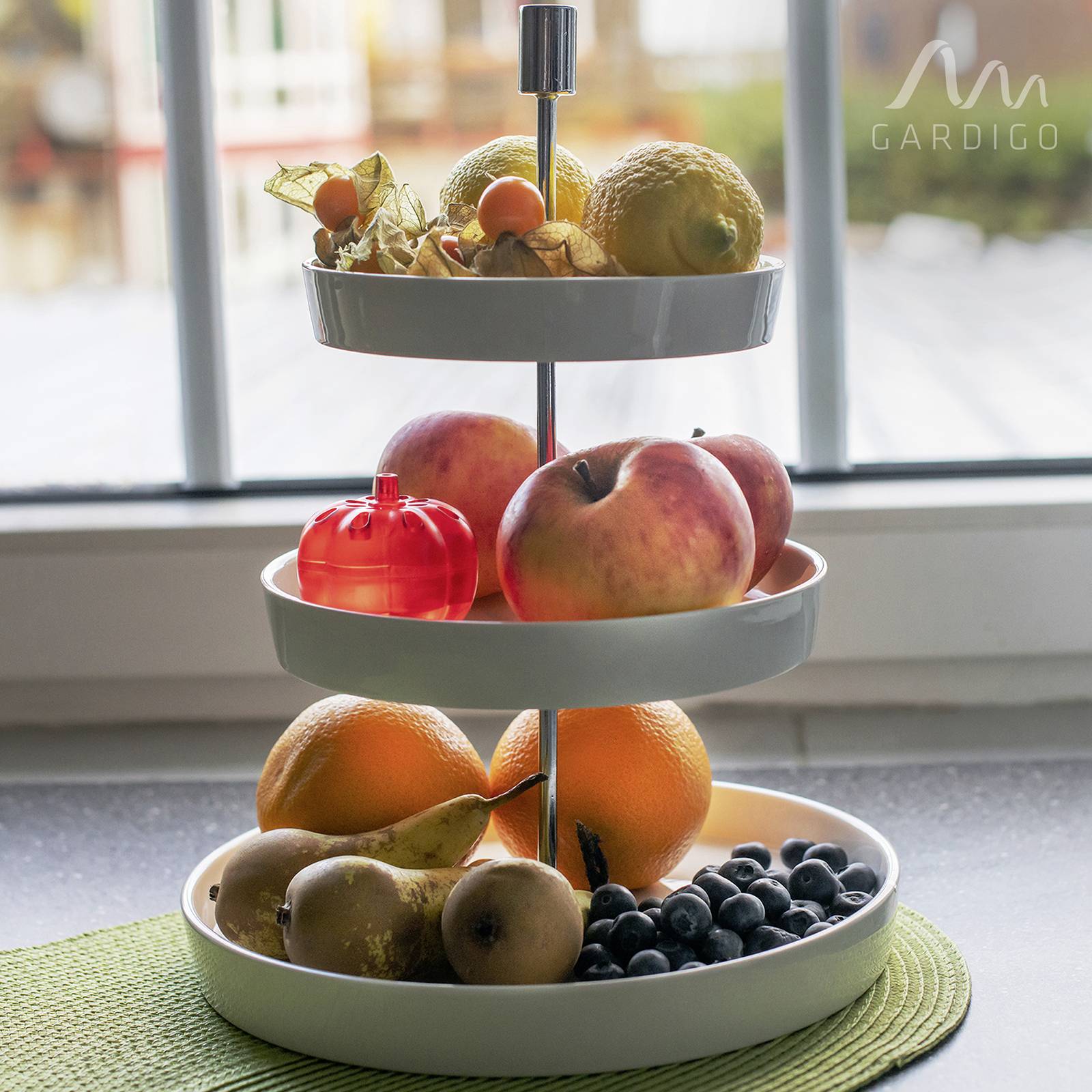 Een drielaags fruitstandaard op een tafel, gevuld met verschillende soorten fruit zoals appels, sinaasappels en bessen, voor een raam.