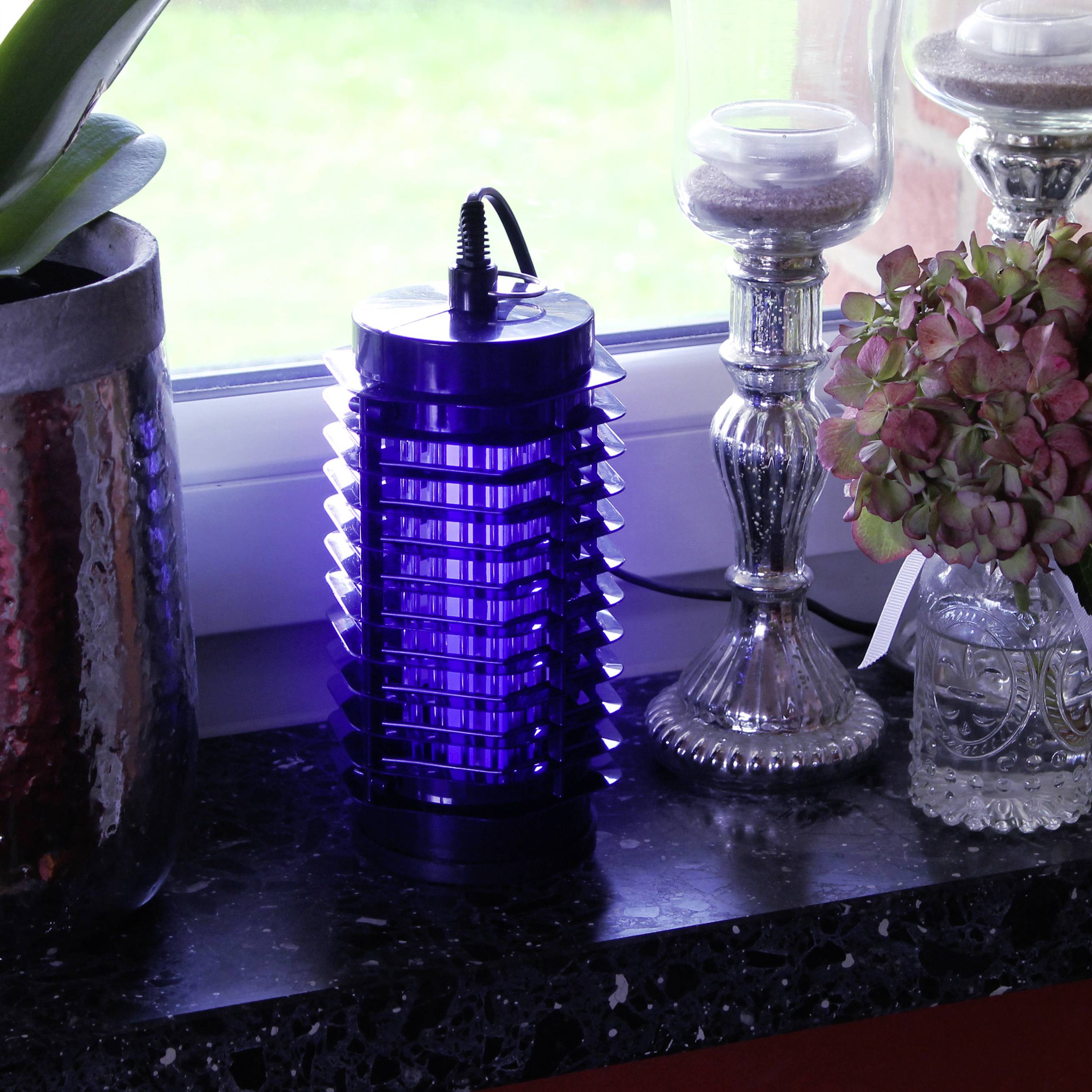 Een blauwe insectenlamp staat op een vensterbank, geflankeerd door planten en kaarsenhouders. Buiten is groene vegetatie zichtbaar.