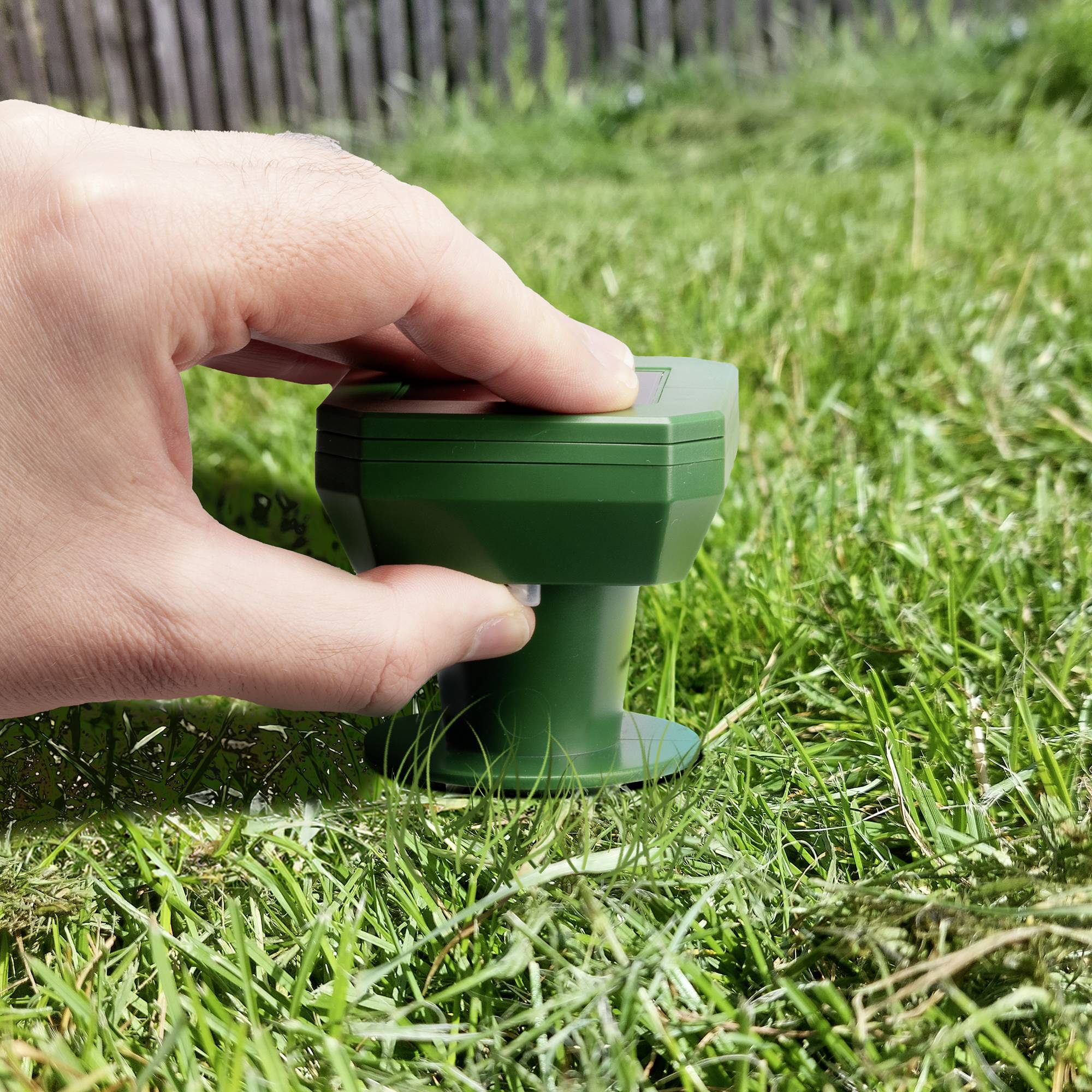 Een hand plaatst een groene sproeier op een groen gazon. Houten hek op de achtergrond.