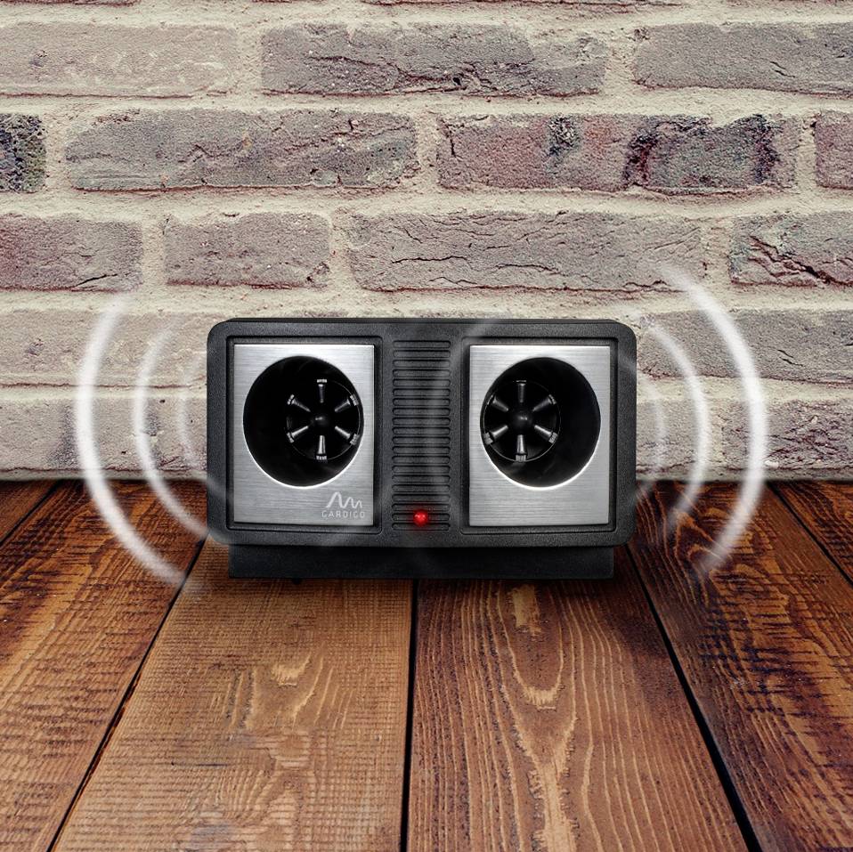 Een elektrisch apparaat met twee luidsprekers en een verlichte weergave staat op een houten vloer voor een bakstenen muur.