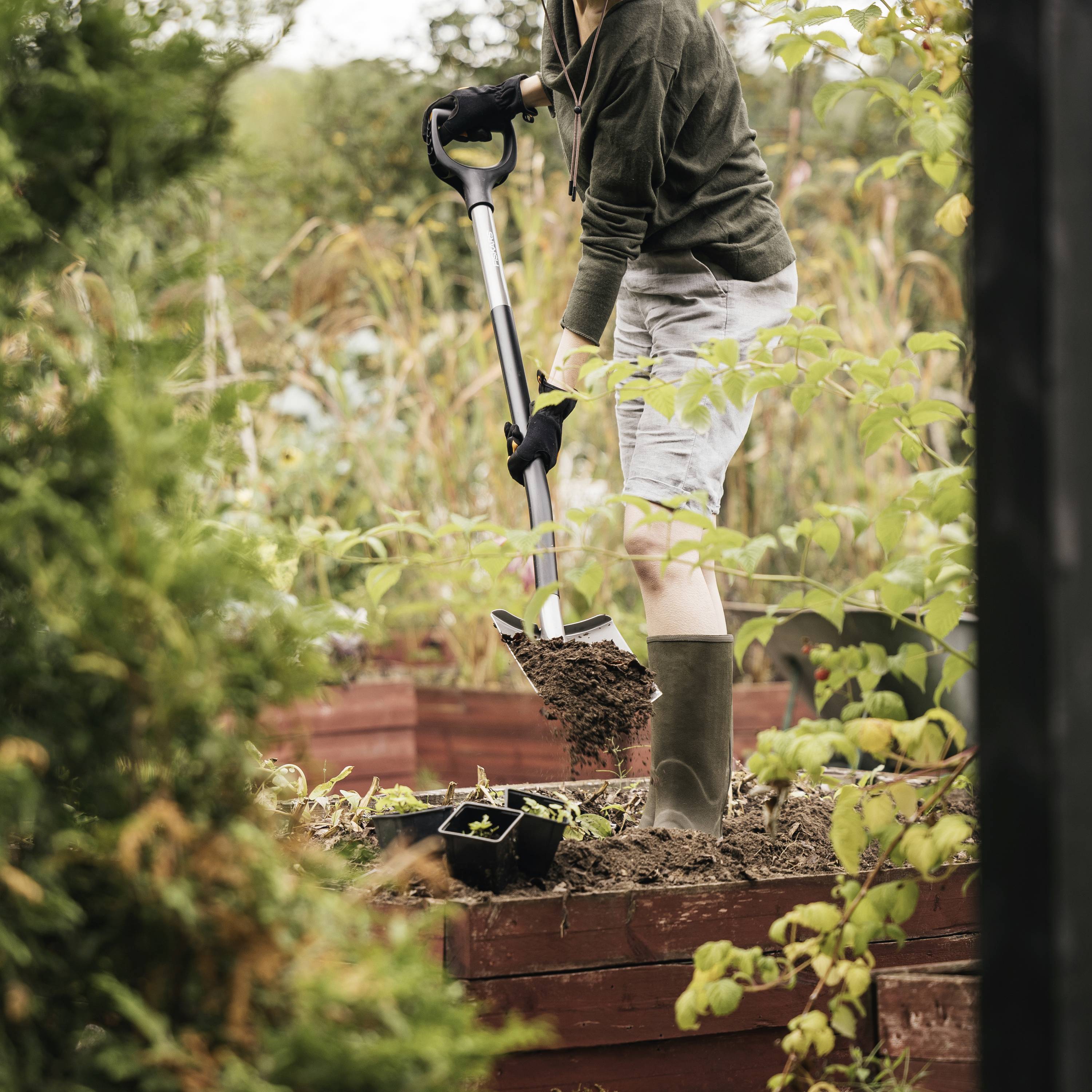 Persoon in de tuin die met een schep de grond omwoelt, omringd door planten.