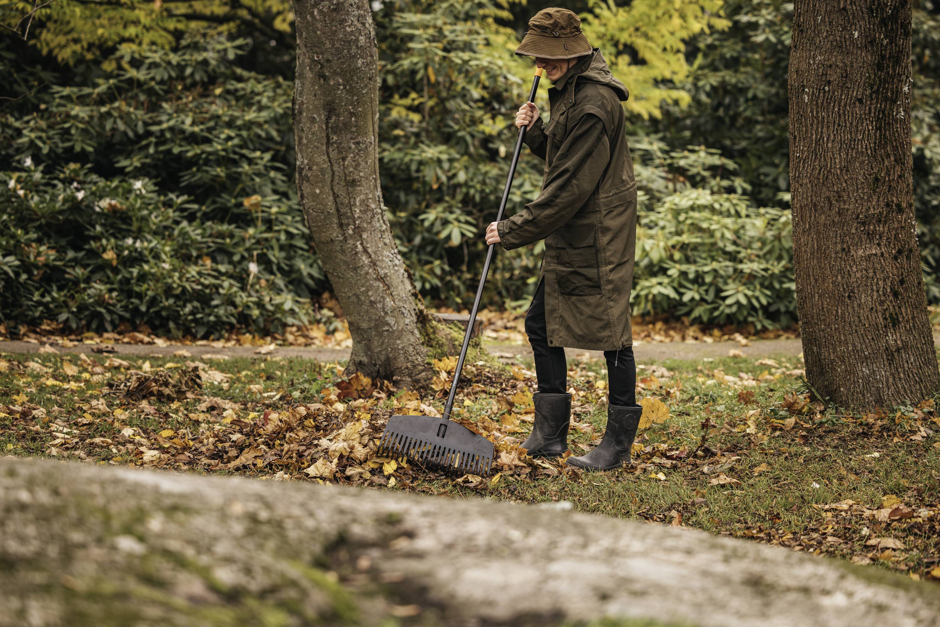Een persoon in groene kleding en rubberen laarzen harkt bladeren in een herfstelijk park. Bomen en bladerhopen zijn op de achtergrond zichtbaar.