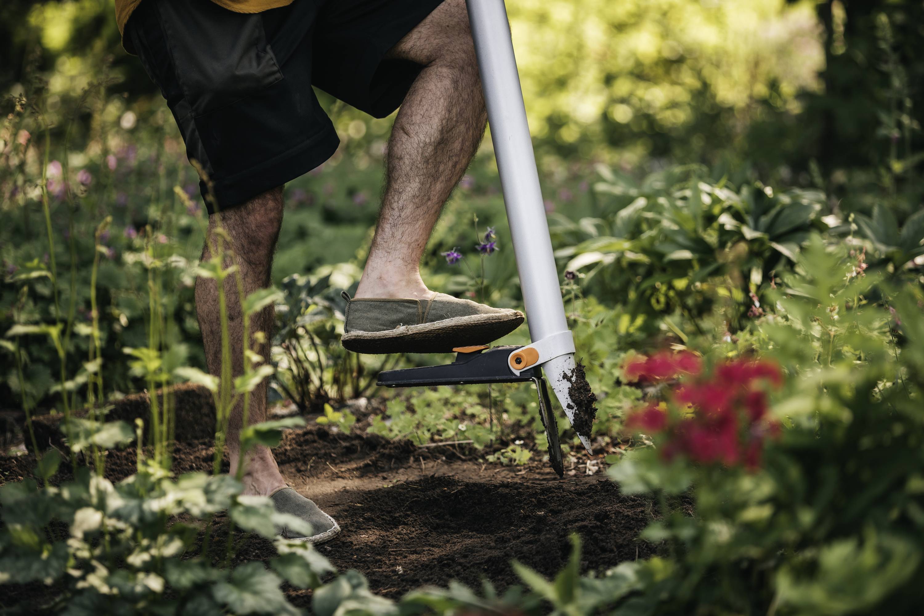 Een persoon graaft met een tuingereedschap in de grond, omringd door groen gebladerte en rode bloemen in een tuin.