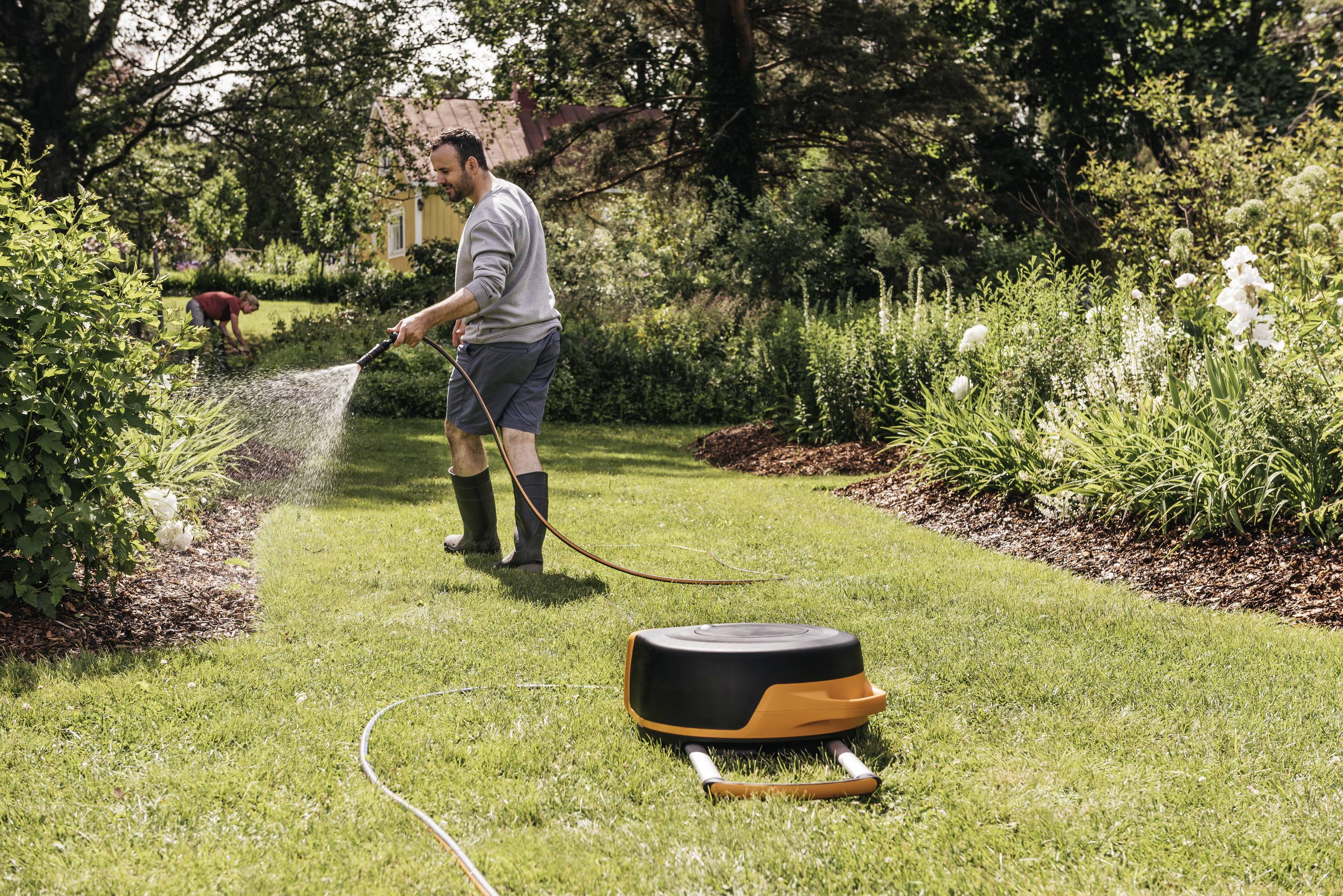 Een man besproeit de tuin met een tuinslang, terwijl een automatische grasmaaier op het gazon staat in de voorgrond.