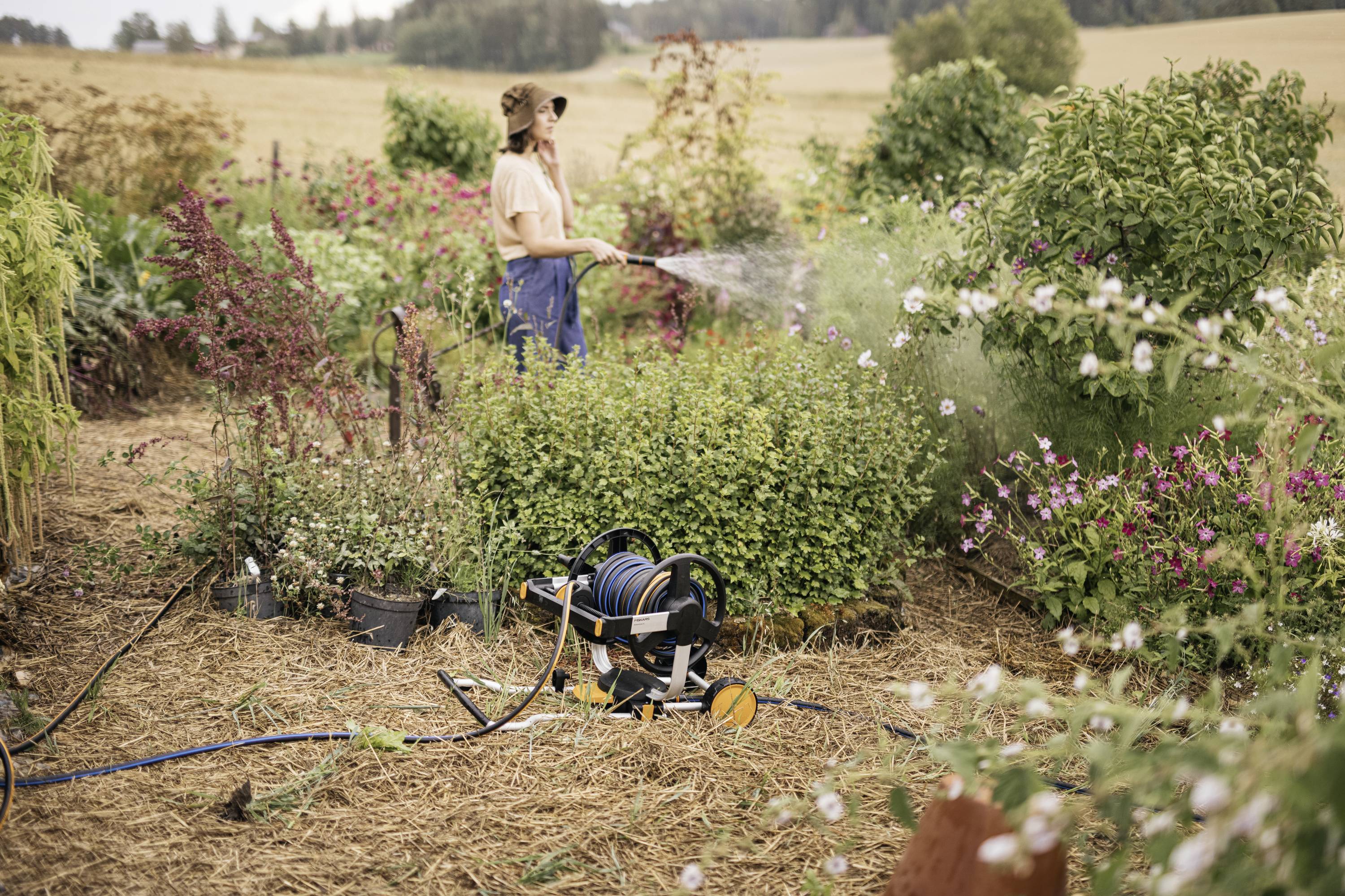 Een persoon begiet planten in de tuin met een tuinslang; omringd door bloeiende struiken. Landelijk landschap op de achtergrond.