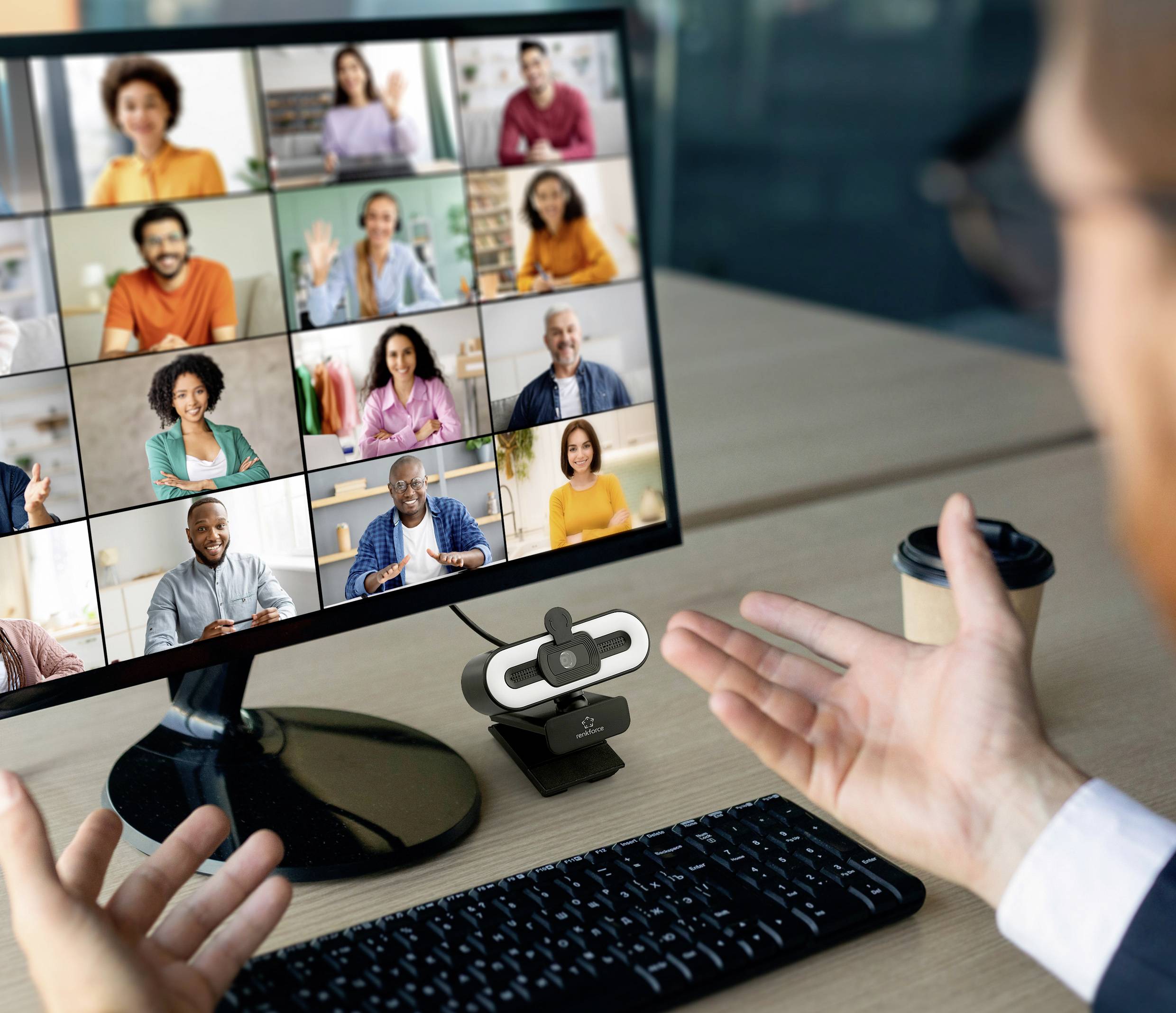 Een persoon zit aan een bureau voor een computerscherm waarop een videogesprek met meerdere deelnemers wordt weergegeven. Handen gebaren expressief.