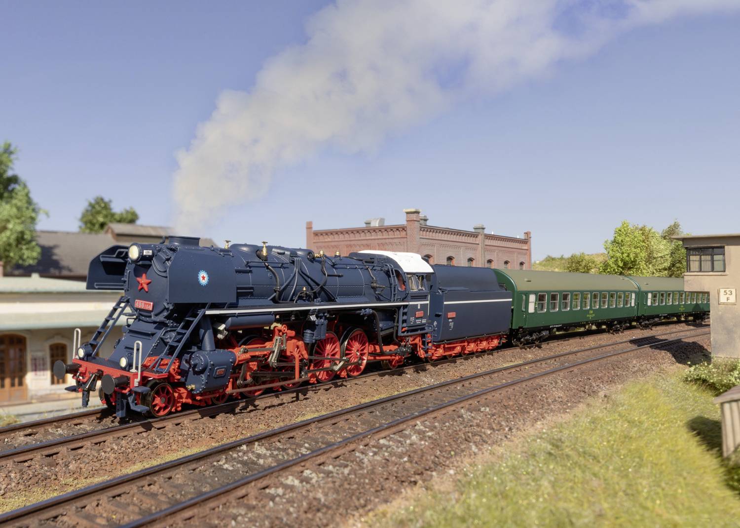 Een blauw-rode stoomlocomotief trekt groene wagons over sporen door een landelijk landschap met bomen en een bakstenen gebouw op de achtergrond.