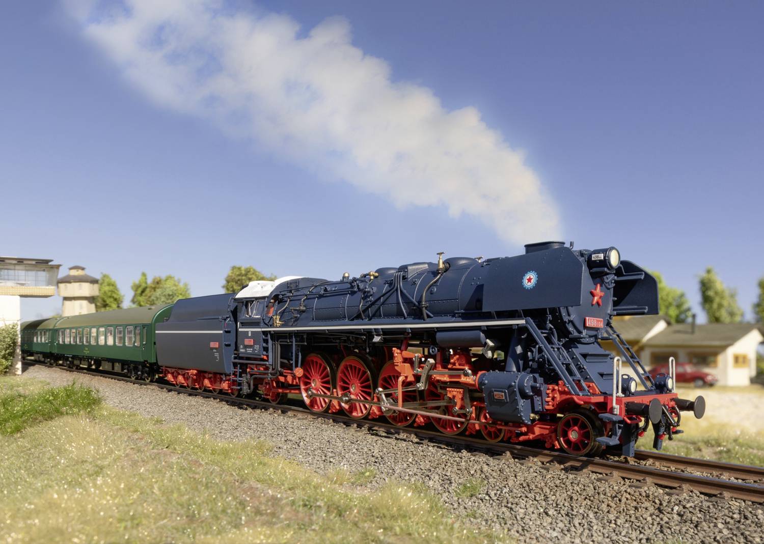Een modelspoordamplocomotief trekt verschillende groene wagons over een spoor door een landelijk landschap onder een heldere hemel.
