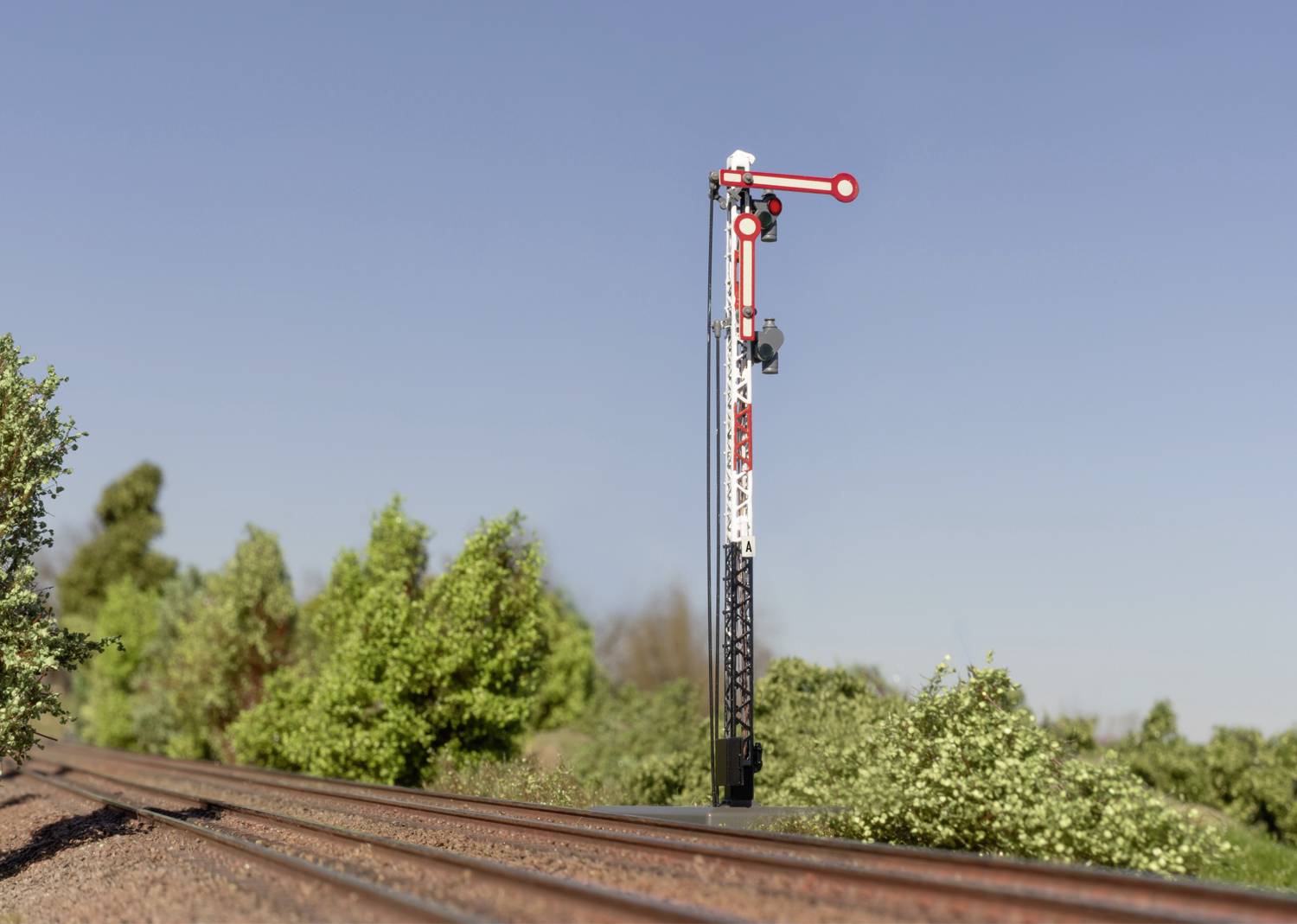 Een mechanisch spoorwegseinsignaal toont 'Stop' aan een spoorrail, omgeven door struiken onder een heldere blauwe lucht.