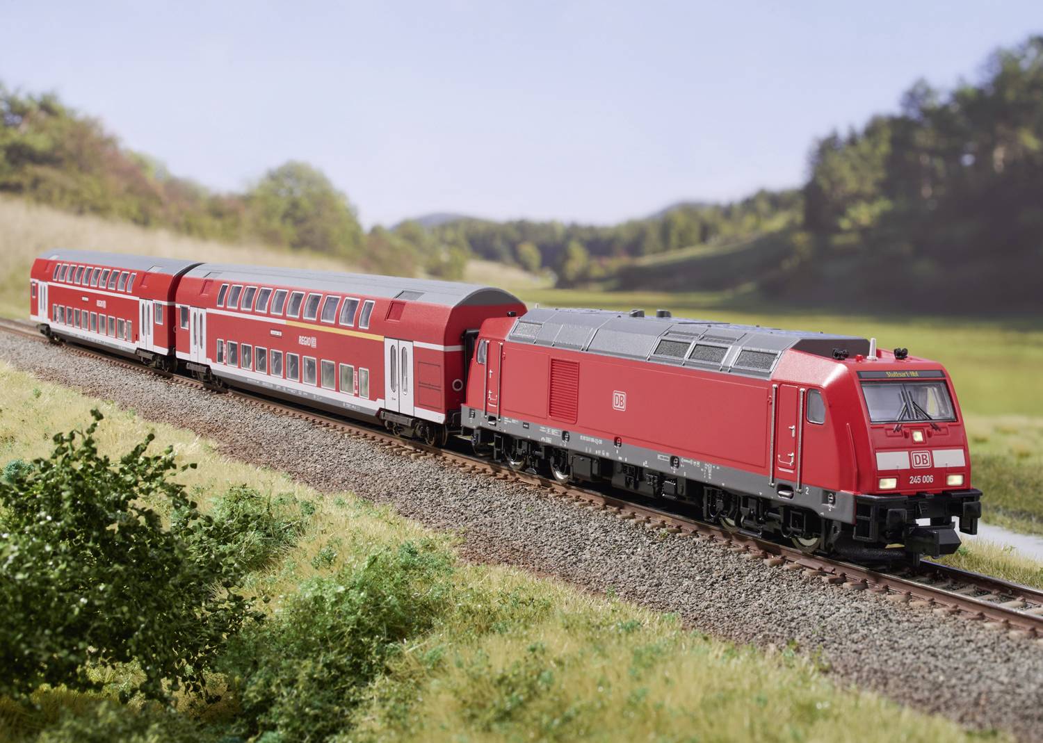 Een rode regionale trein rijdt door een groene landschap met bomen op de achtergrond. Een heldere blauwe lucht wijst op mooi weer.