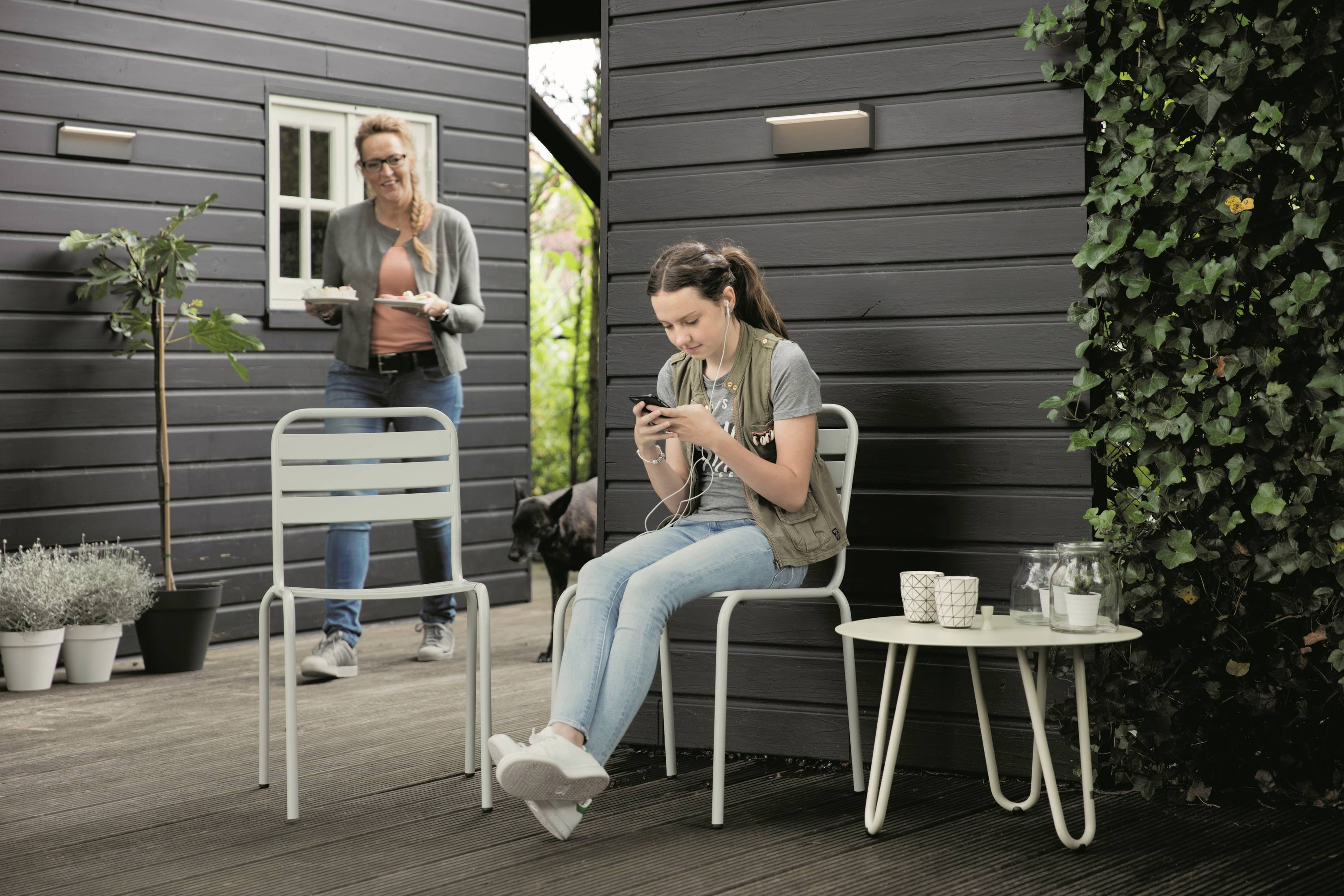 Een vrouw zit buiten op een stoel en kijkt naar haar telefoon. Een andere persoon komt met een dienblad uit een gebouw.