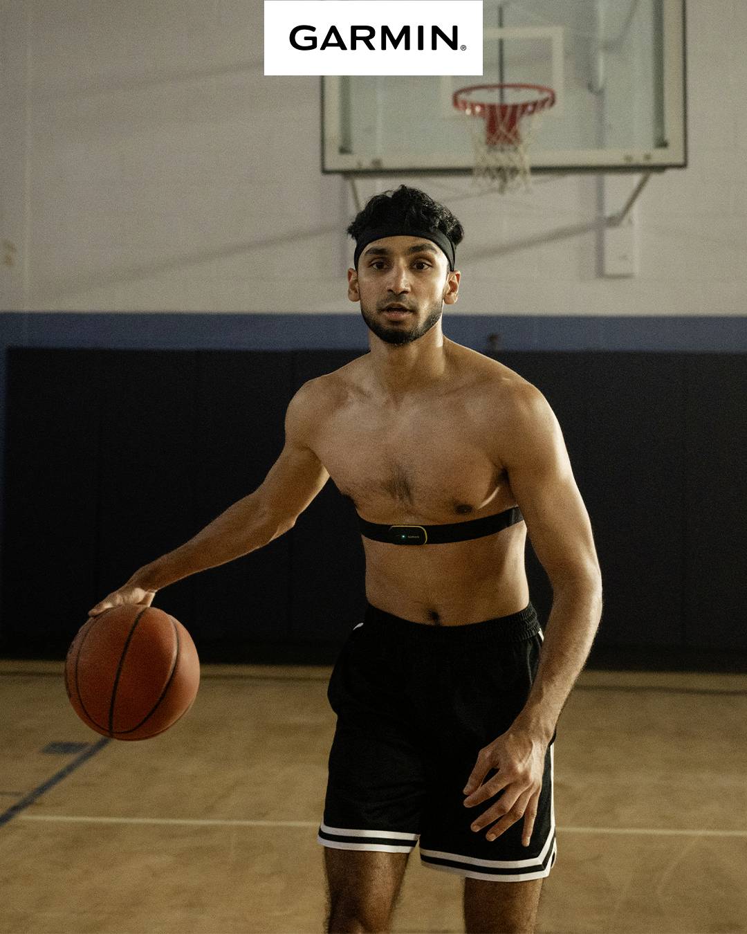 Een man met een basketbal draagt een fitness-borstband in een sporthal. Op de achtergrond is een basketbalring te zien. Bovenaan staat 'GARMIN'.