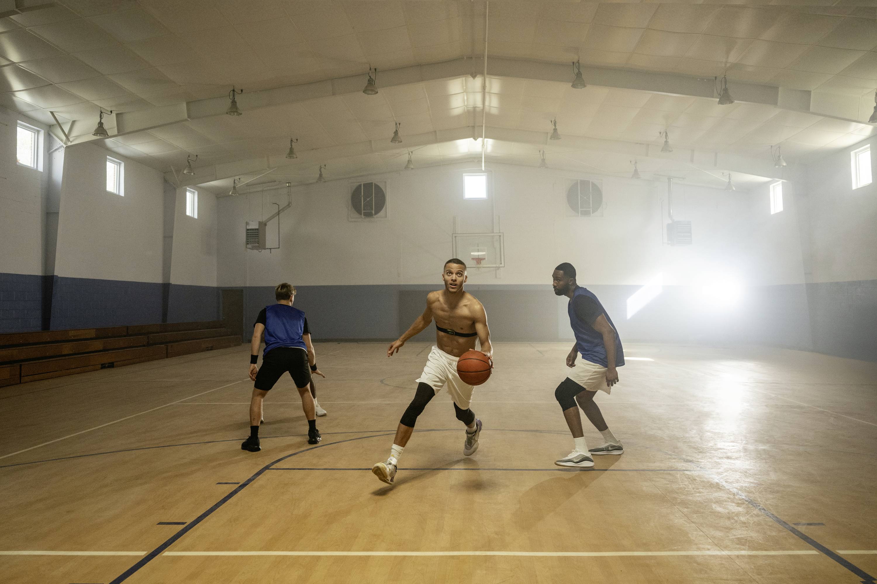 Drie mannen spelen basketbal in een sporthal. Een speler dribbelt met de bal, terwijl de andere twee hem verdedigen.