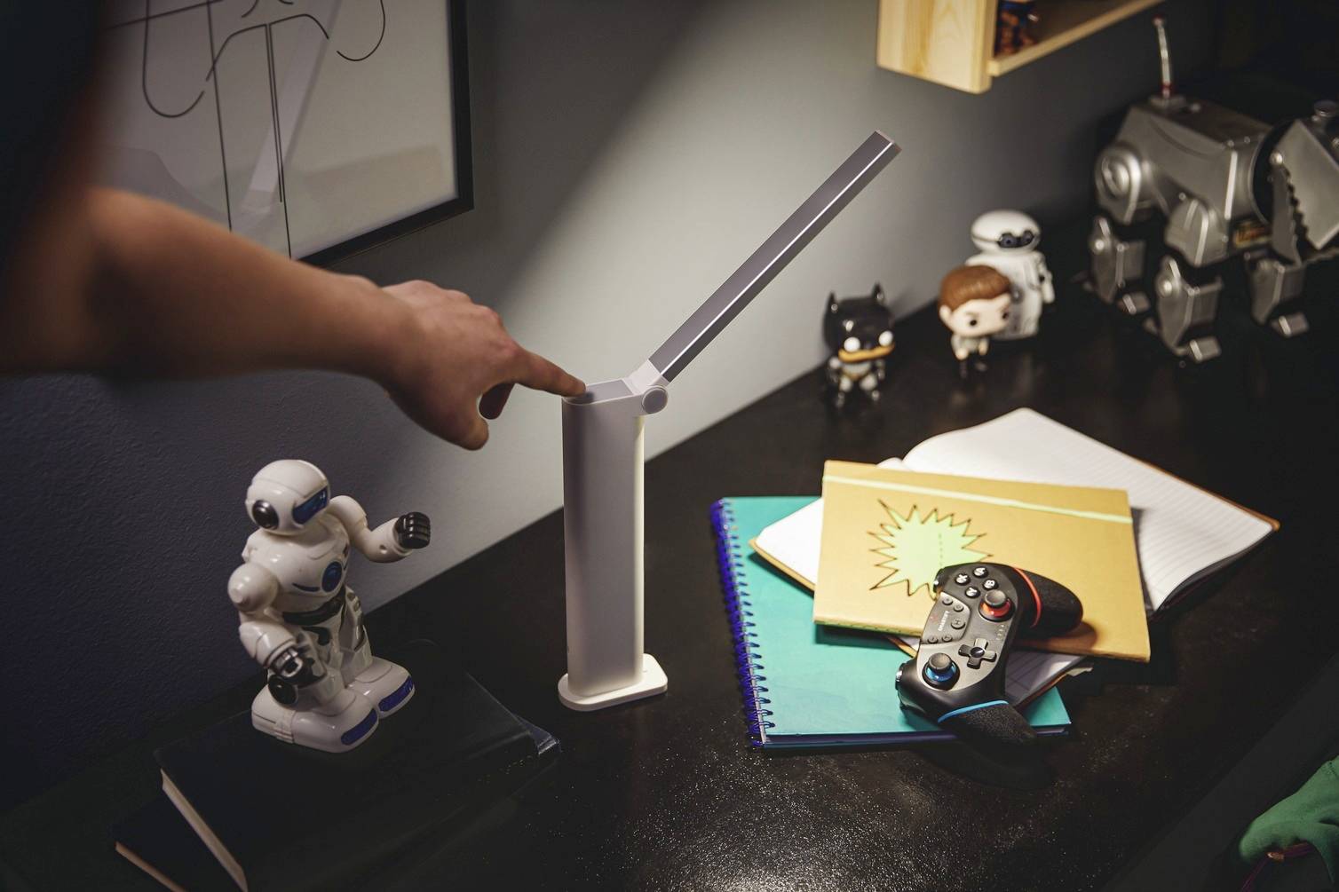 Bureau met notitieboeken, gamecontroller, figuren en verstelbare lamp; persoon wijst naar de lampknop.