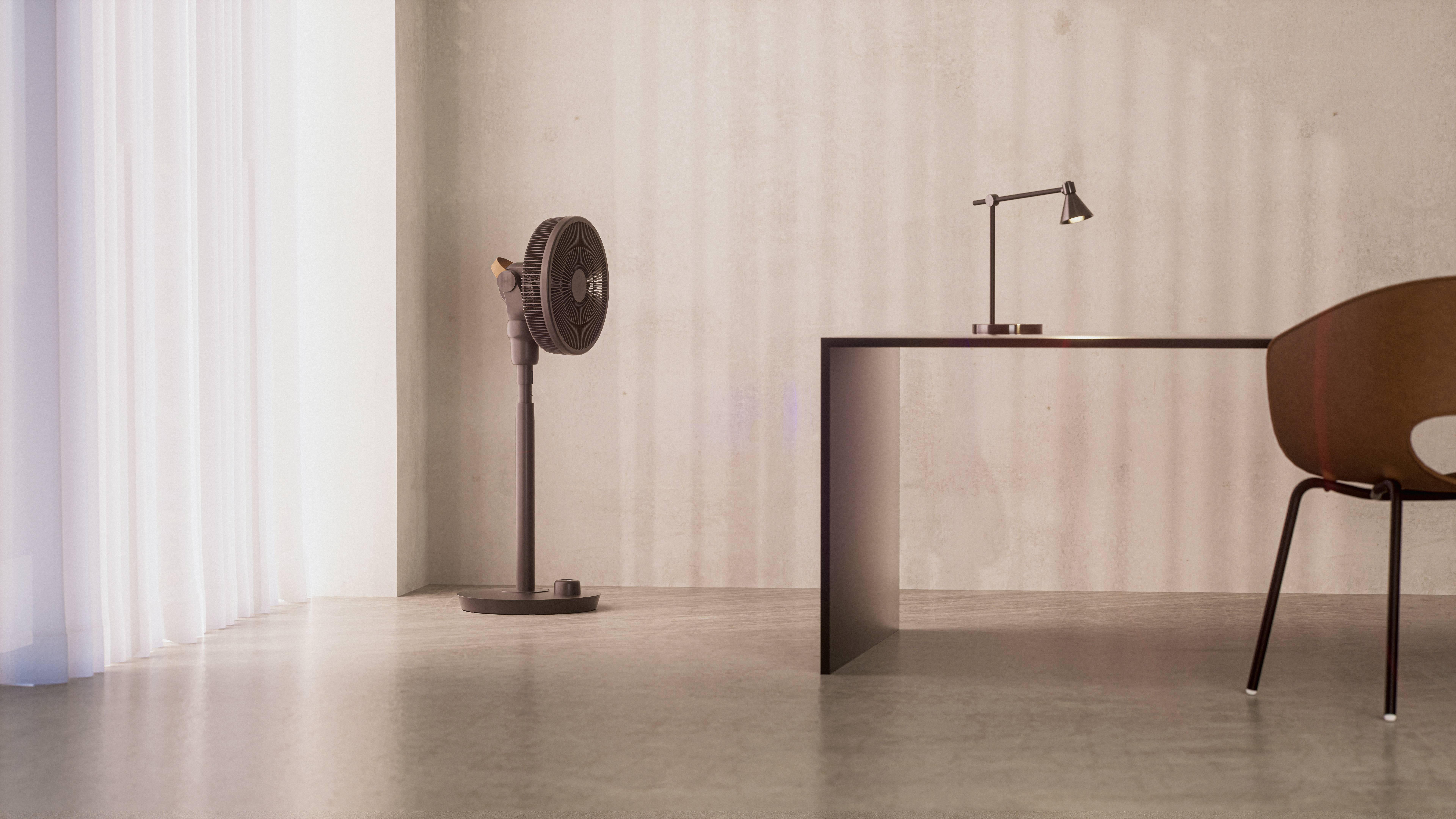 Minimalistische ruimte met bureau, lamp, stoel en staande ventilator. Licht valt door een gordijn op de betonnen vloer.