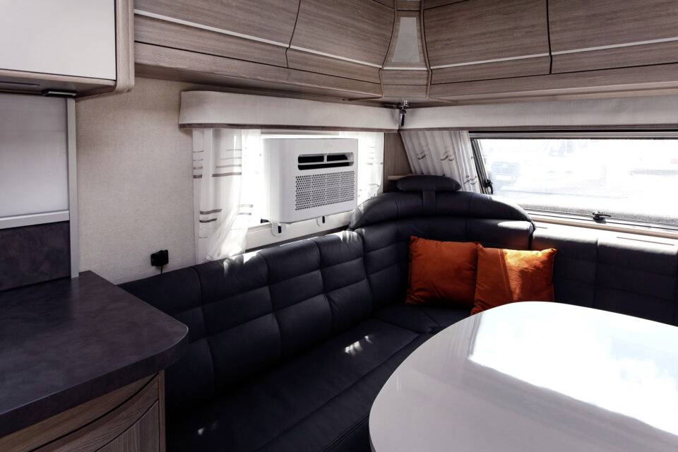 Interieur van een camper met zwarte leren bank, oranje kussens en een ronde tafel. Een raam laat wat daglicht binnen.