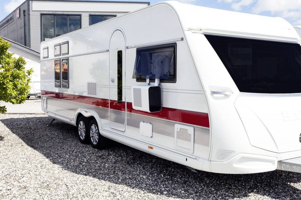 Een witte caravan met rode strepen staat op een kiezelplaats voor een gebouw. Heldere hemel en zonnig weer.