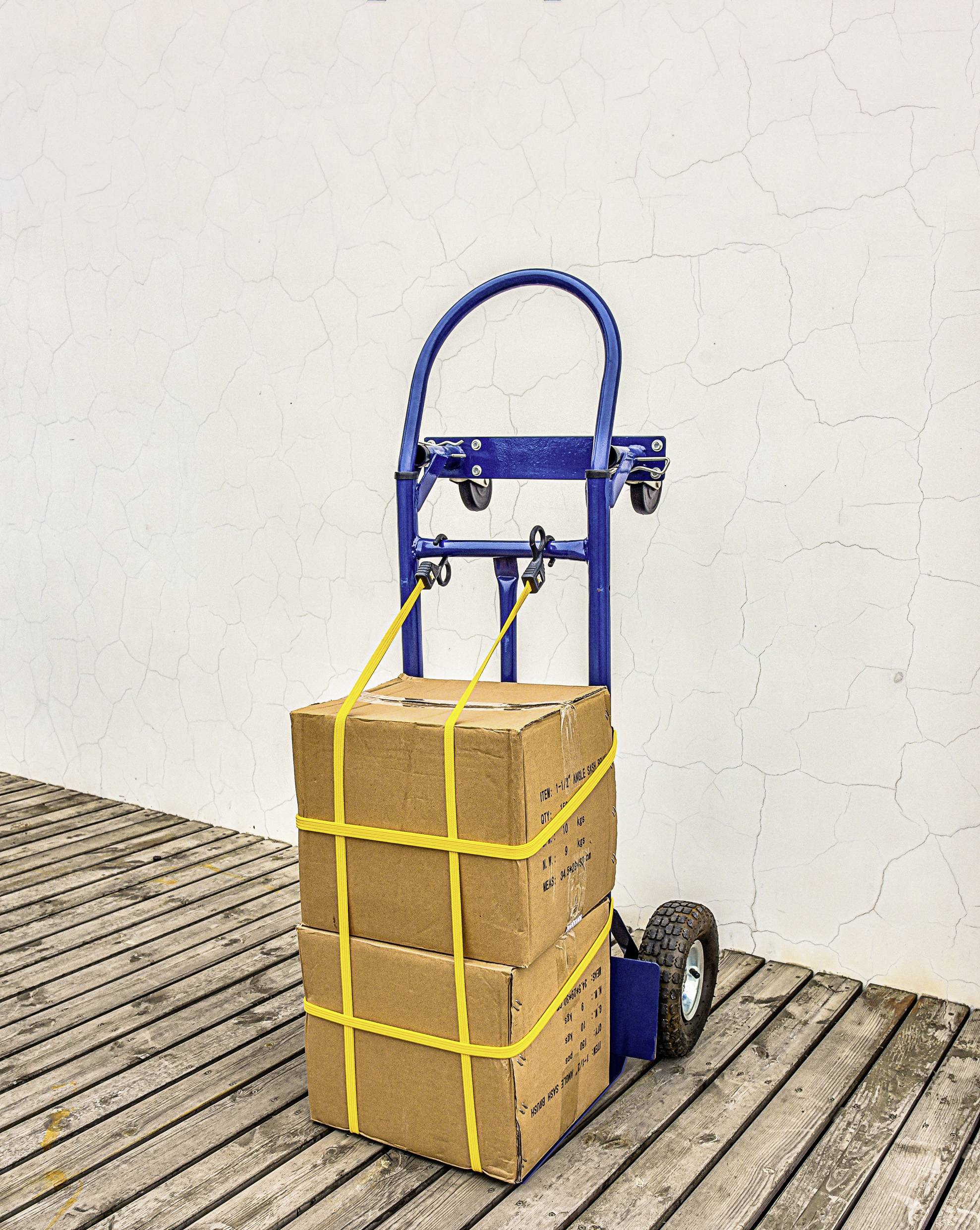 Een blauwe steekwagen op een houten terras draagt twee gestapelde kartonnen dozen die zijn vastgemaakt met gele riemen, geplaatst tegen een witte gestructureerde muur.