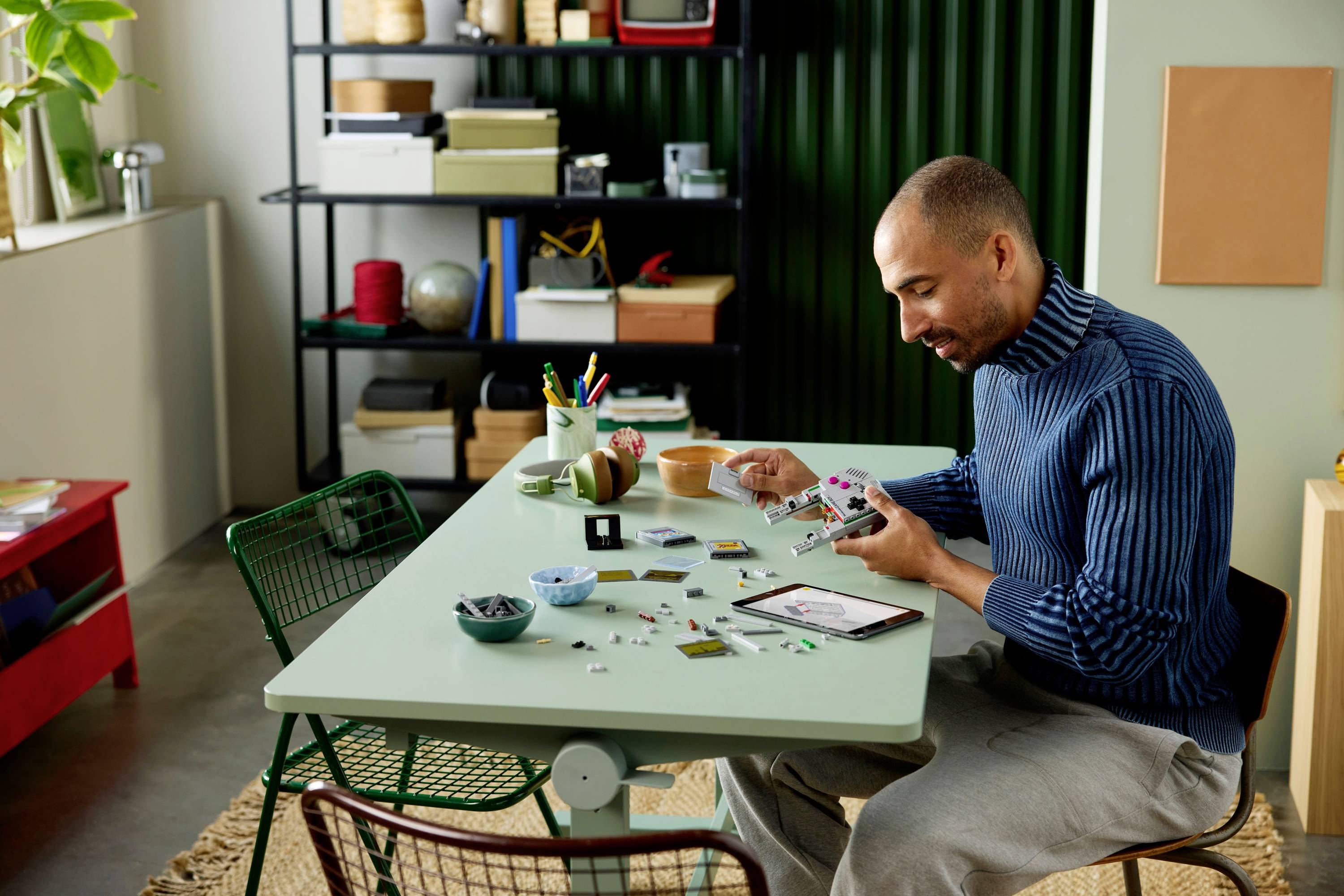 Man zit aan een tafel kleine modelbouwonderdelen in elkaar te zetten, met verschillende onderdelen verspreid voor hem. Boekenkasten met dozen en decoratie-items op de achtergrond.