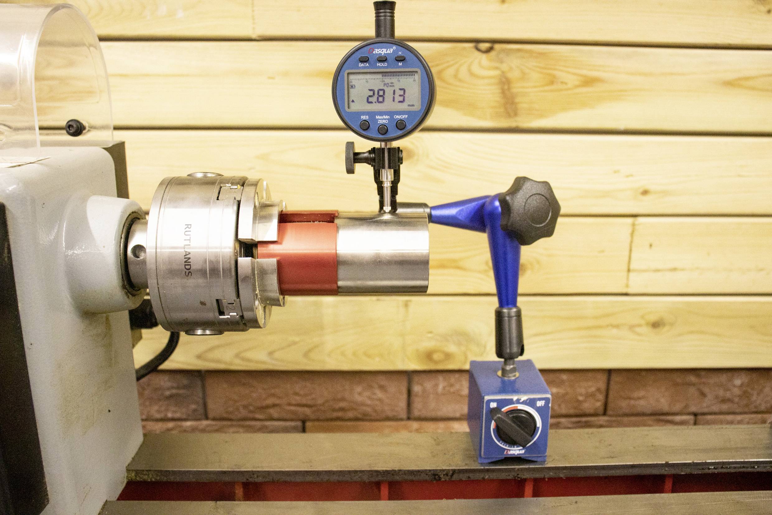 Een meetklok meet de diameter van een metalen cilinder die in een draaibank is geklemd, waarbij '28,13' wordt weergegeven tegen een houten wandachtergrond.