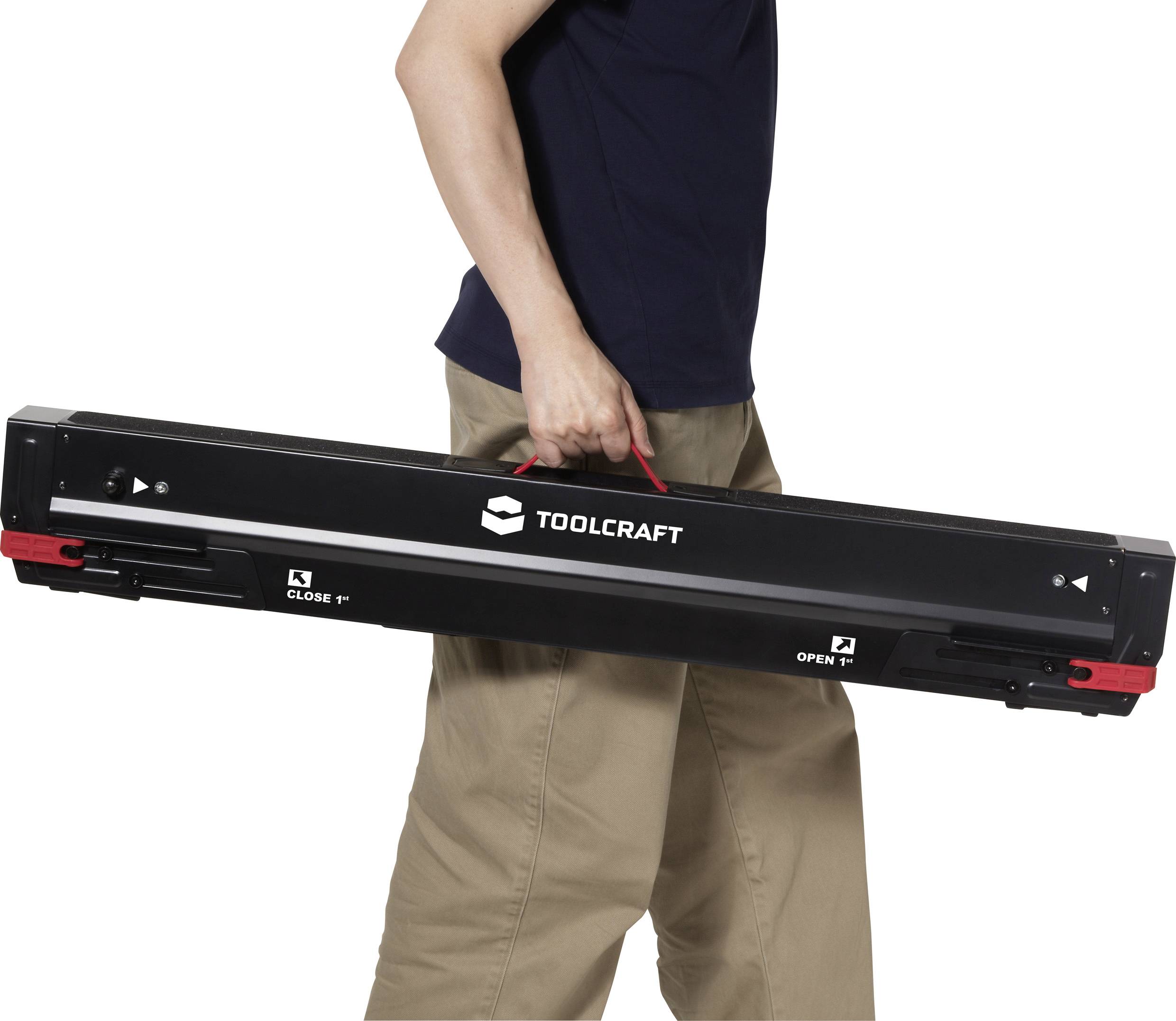Een persoon in beige broek en een donker shirt die een lange zwarte Toolcraft gereedschapskist draagt met een handvat, waarbij 'Sluiten' en 'Openen' instructies worden getoond.