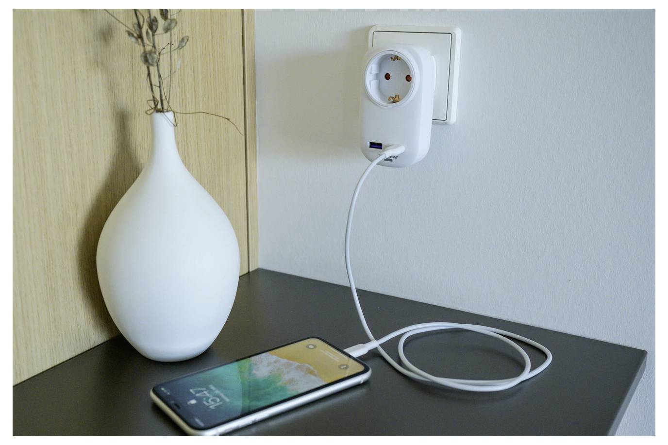 Een smartphone wordt opgeladen op een tafelblad naast een wandstekkerdoos, met een opgerold wit snoer dat ingesteekt is en een witte vaas op de achtergrond.