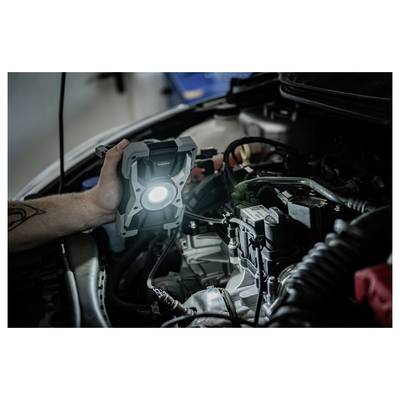 Een persoon houdt een verlichte werkverlichting in de motorruimte van een auto vast, waarbij deze de motor inspecteert of reparaert.