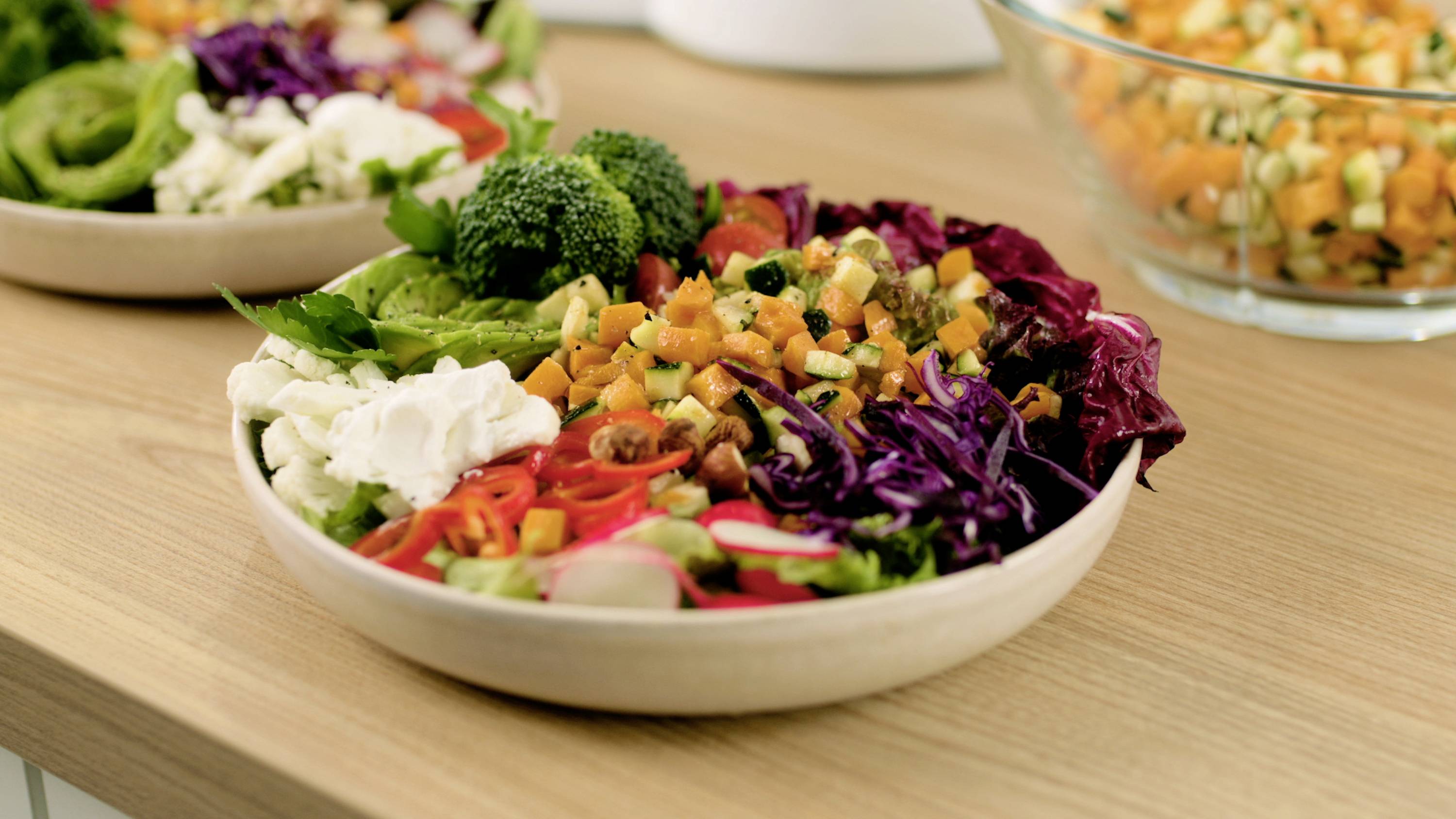 Een kom met kleurrijke gemengde salade met broccoli, sla, radijsjes, rode kool, gewürfelde oranje groenten en komkommers op een houten tafel.