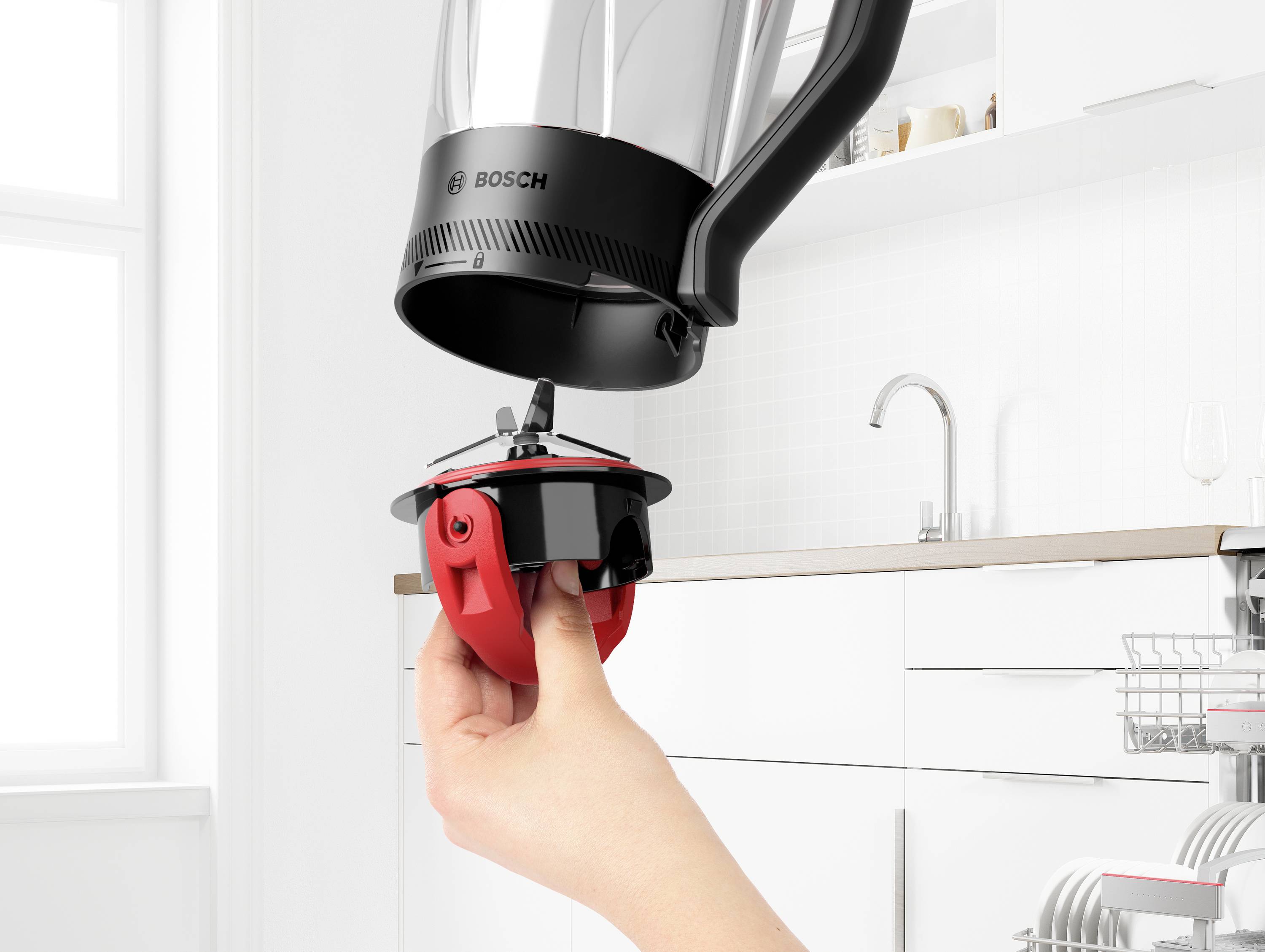 Een hand houdt een rode gardeelmenger vast onder een Bosch keukenmachine, klaar om te worden bevestigd. De keuken heeft moderne, witte kasten.
