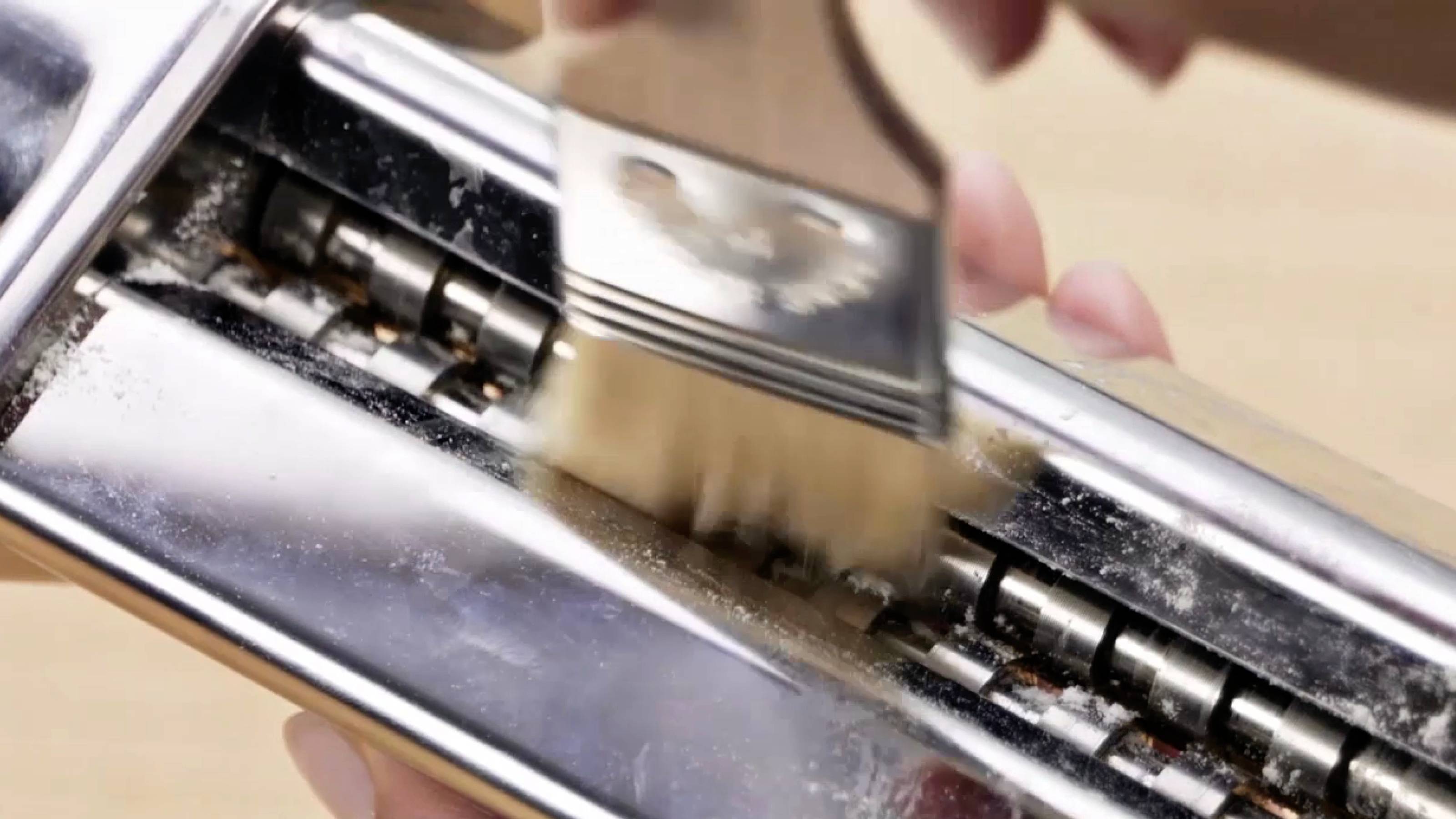 Een persoon gebruikt een borstel om restanten te verwijderen van een metalen pastacutter, waarbij de nadruk ligt op de gewrichten en messen.