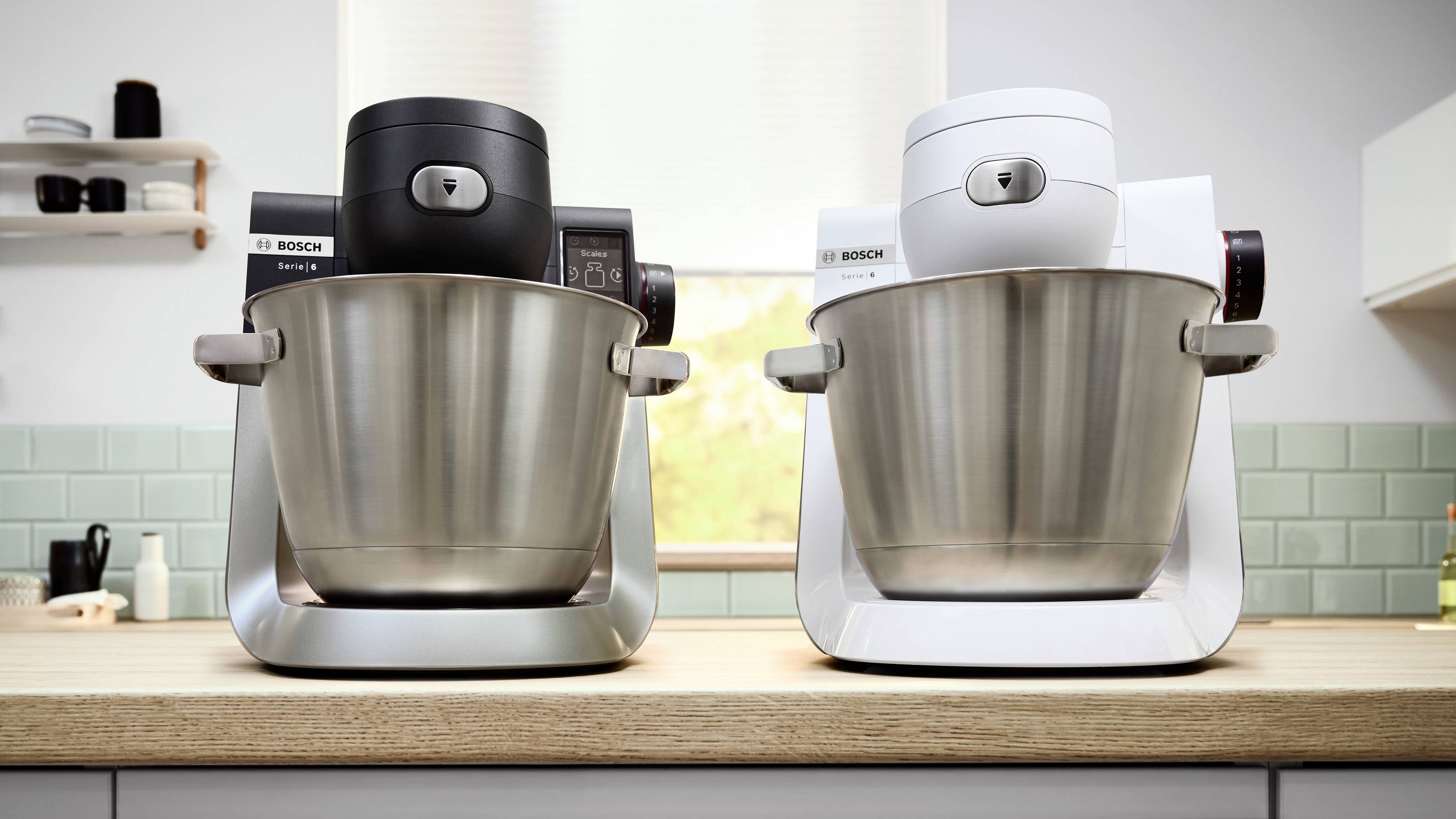 'Twee Bosch keukenmixers, één zwart en één wit, staan op een houten aanrecht in een keuken met een raam en een betegelde achterwand.'