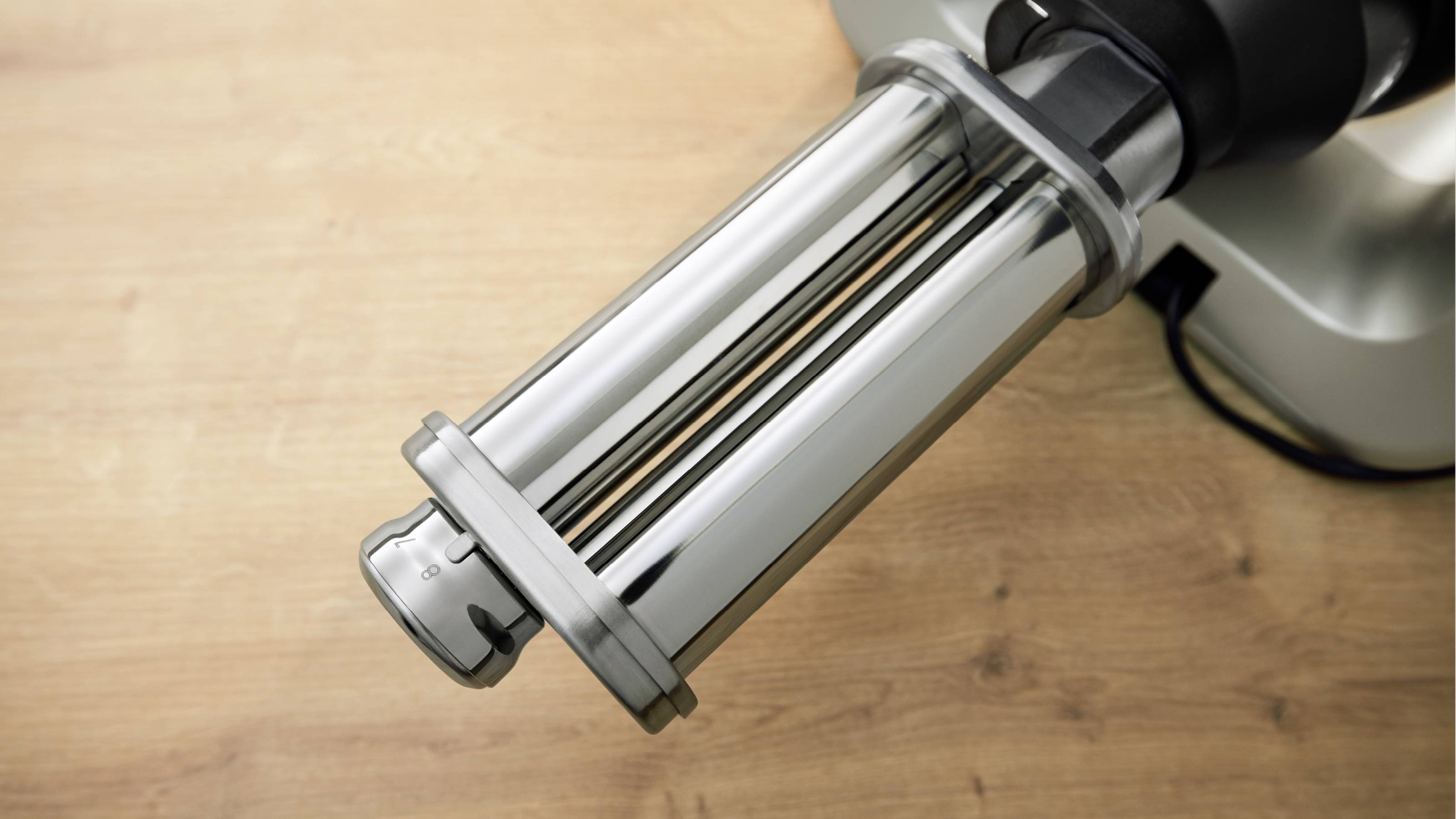 Een metalen pastaroller-accessoire op een keukenmachine, geplaatst op een houten aanrecht, gebruikt voor het uitrollen van pastadeeg.