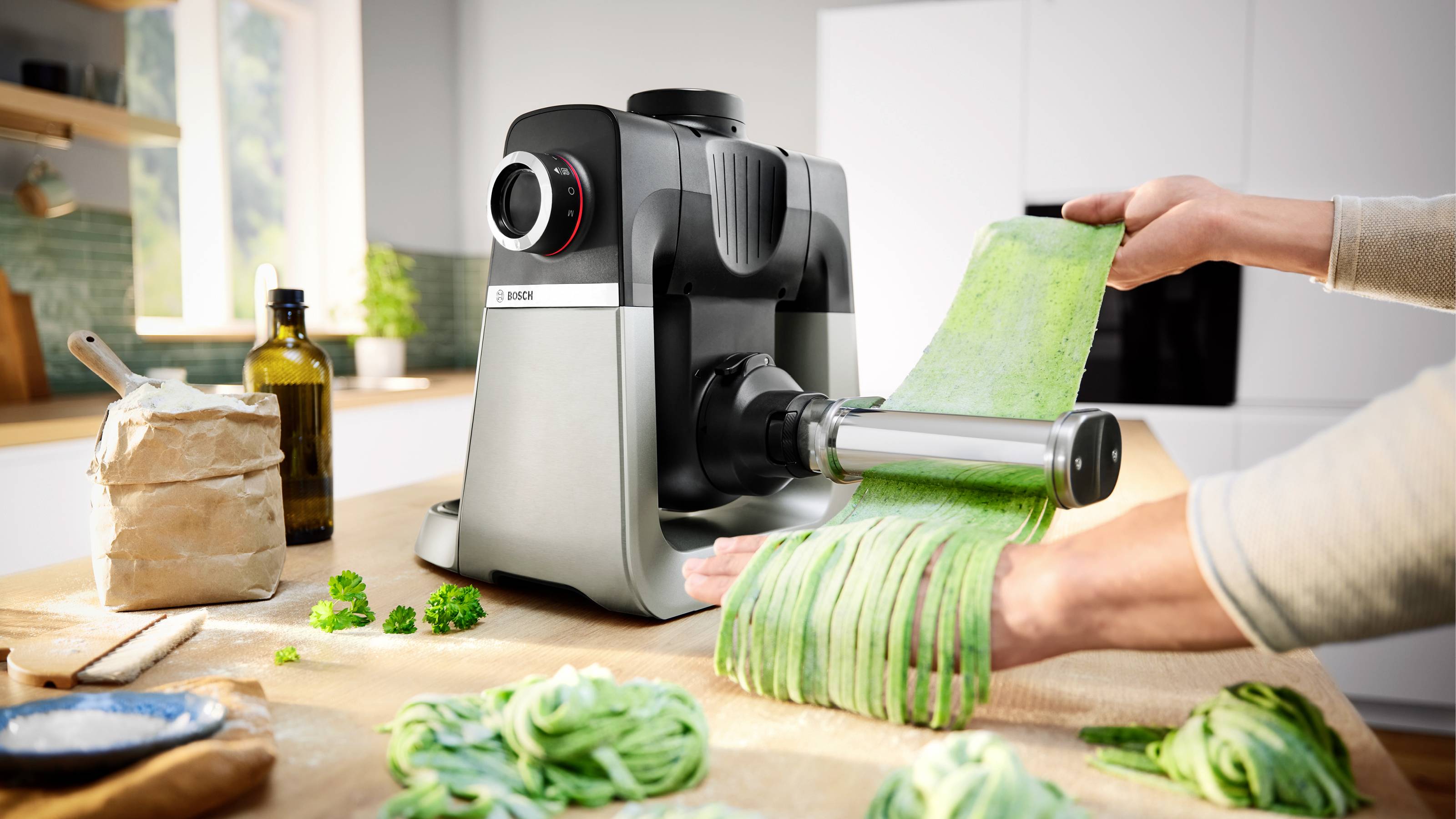 Persoon die verse pasta maakt met een pastamachine in een keuken, omringd door ingrediënten zoals bloem en verse kruiden.