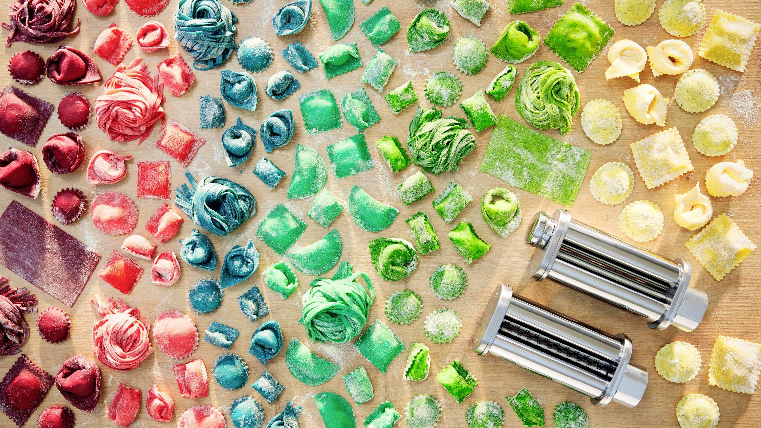 Een verscheidenheid aan meerkleurige pasta in verschillende vormen, waaronder ravioli en fettuccine, gerangschikt in secties per kleur met twee pastasnijders in de buurt.