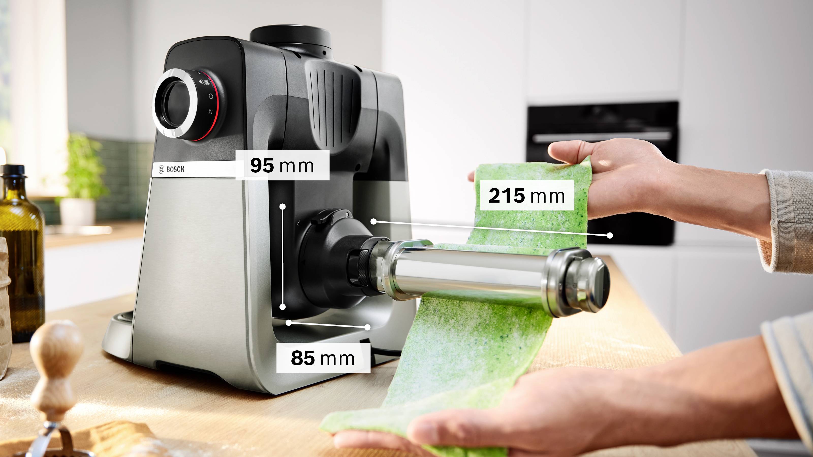 Een pastamachine staat in gebruik op een aanrecht, met een basis van 95 mm en 85 mm. De pastarol is 215 mm breed. Twee handen geleiden groen pastadeeg doorheen het toestel.