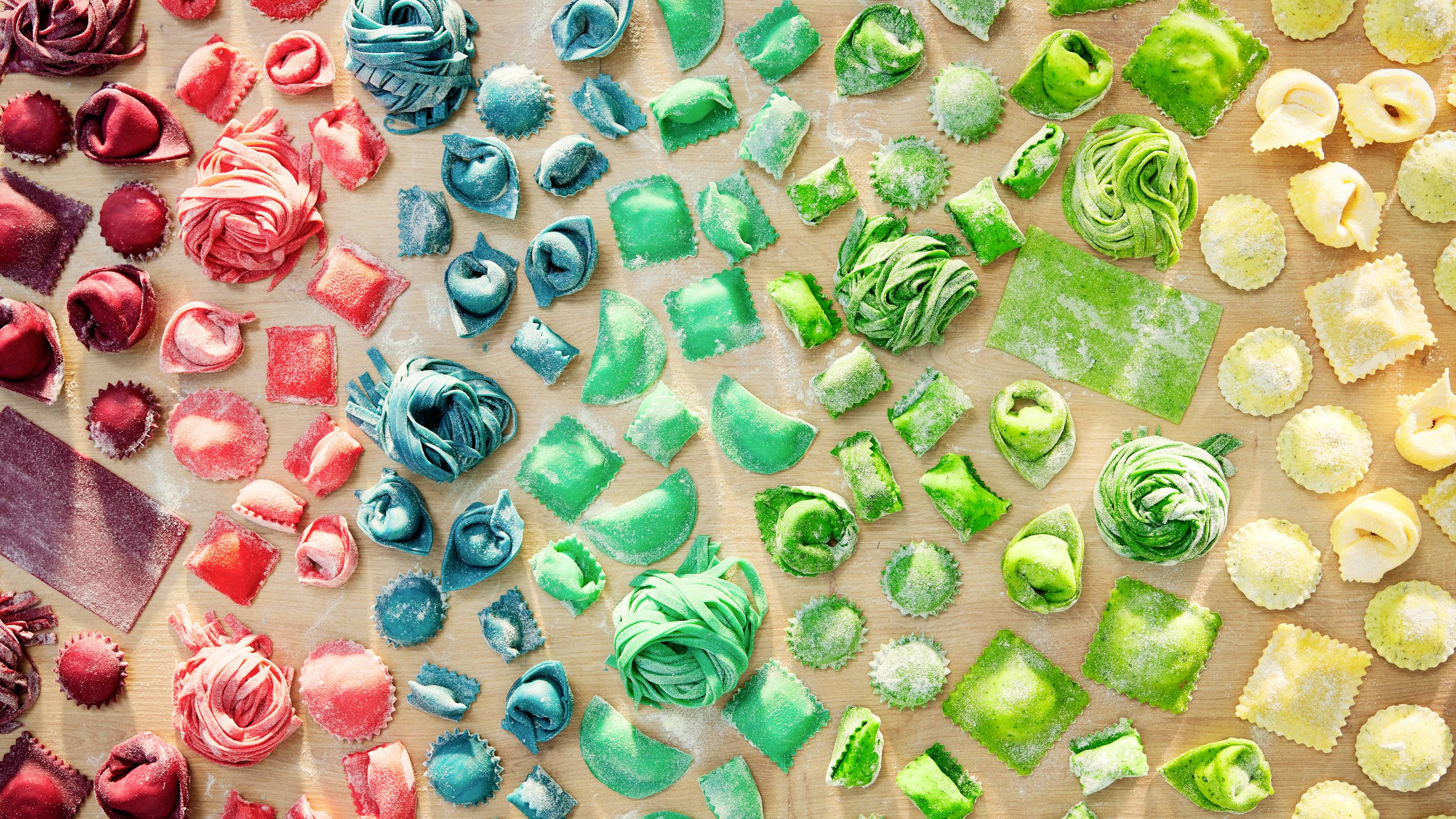 Een verscheidenheid aan kleurrijke handgemaakte pasta, met rode, blauwe en groene tinten, gerangschikt in een gradiëntpatroon op een houten oppervlak.