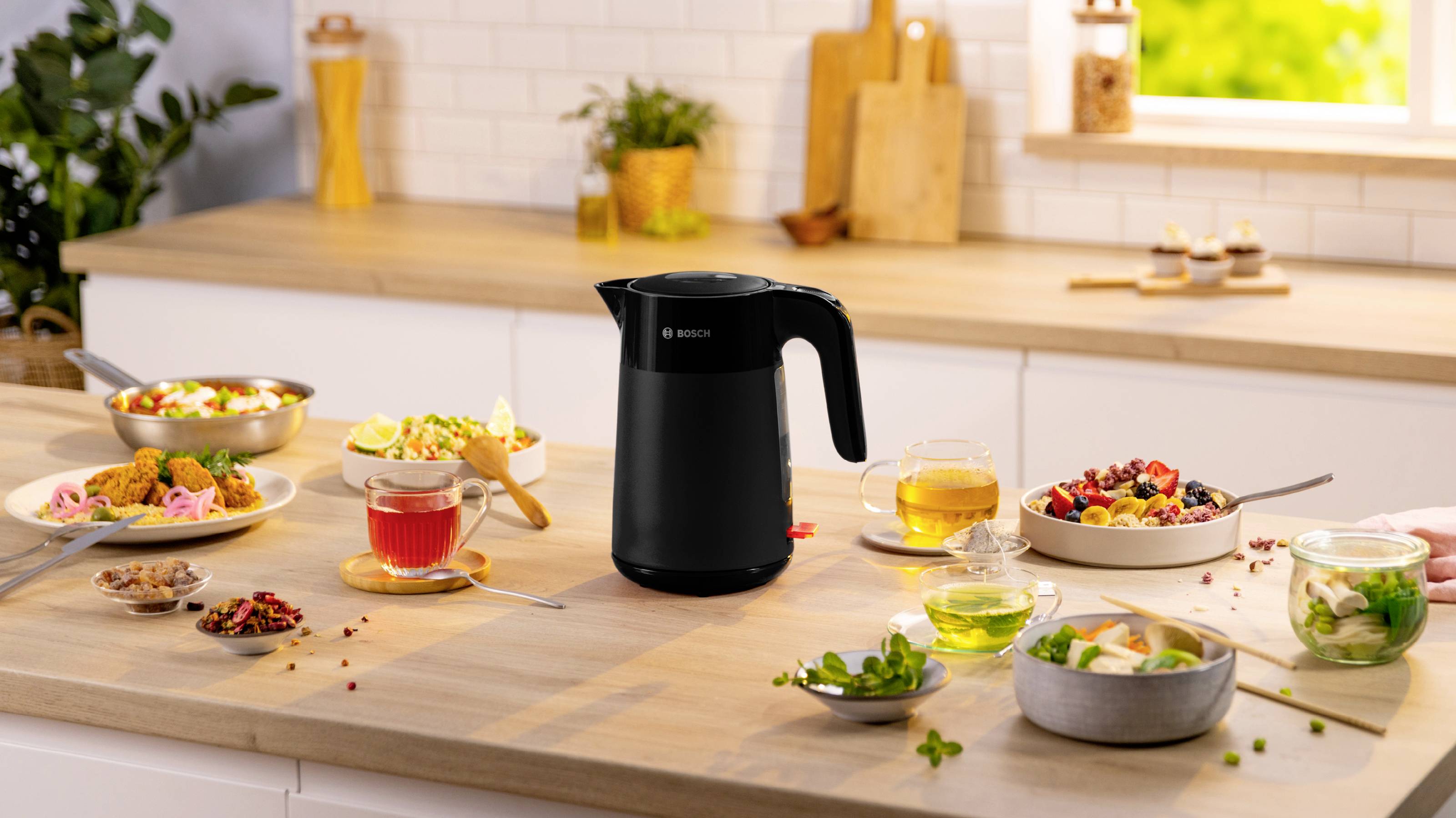 Een zwarte elektrische waterkoker staat op een aanrecht, omringd door kommen en kopjes met kleurrijke salades, thee en ingrediënten, met een lichte, zonnige keukenachtergrond.
