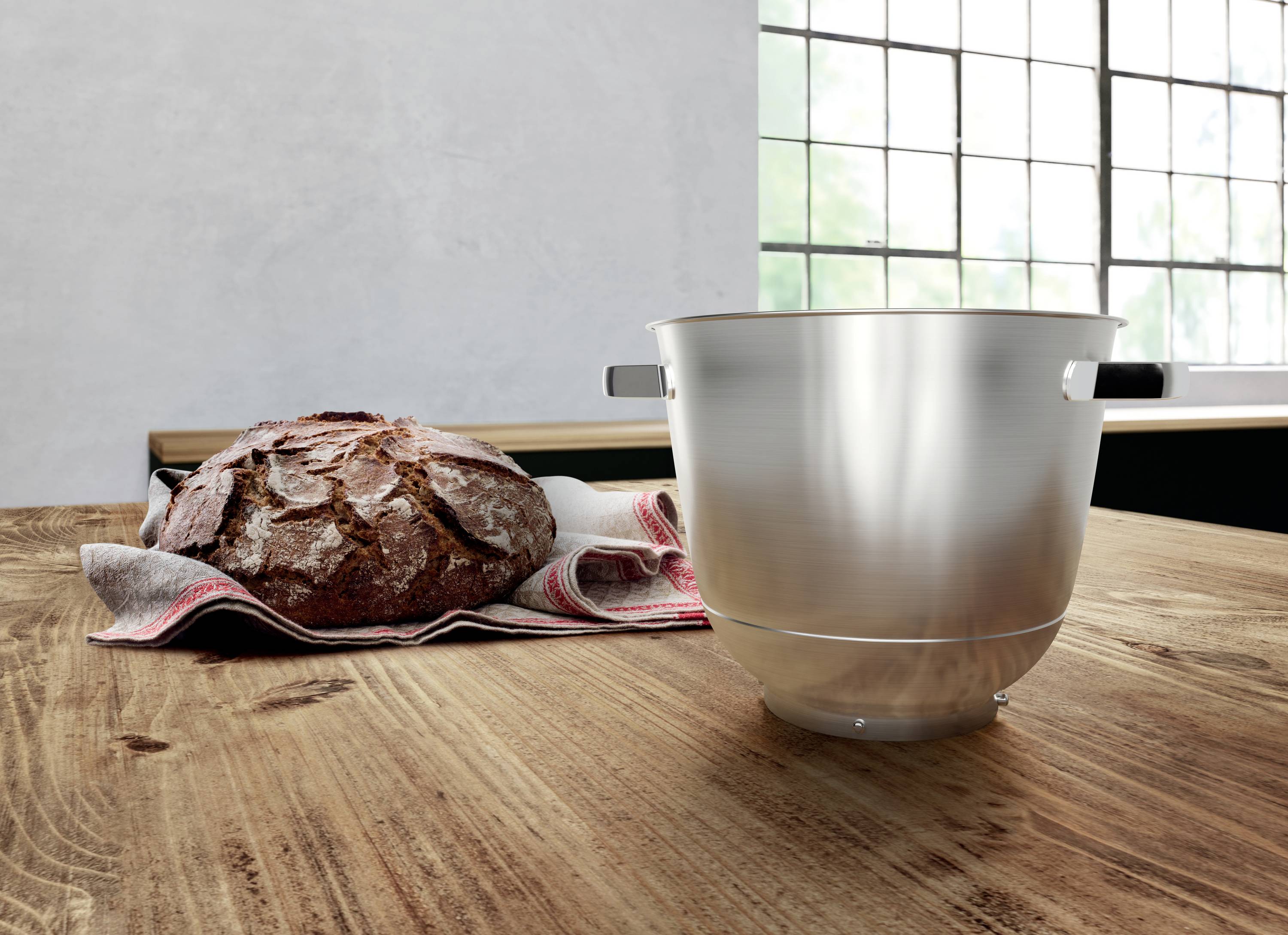 Een rustiek brood ligt op een houten tafel naast een glimmende metalen mengkom, in een keuken met grote ramen.