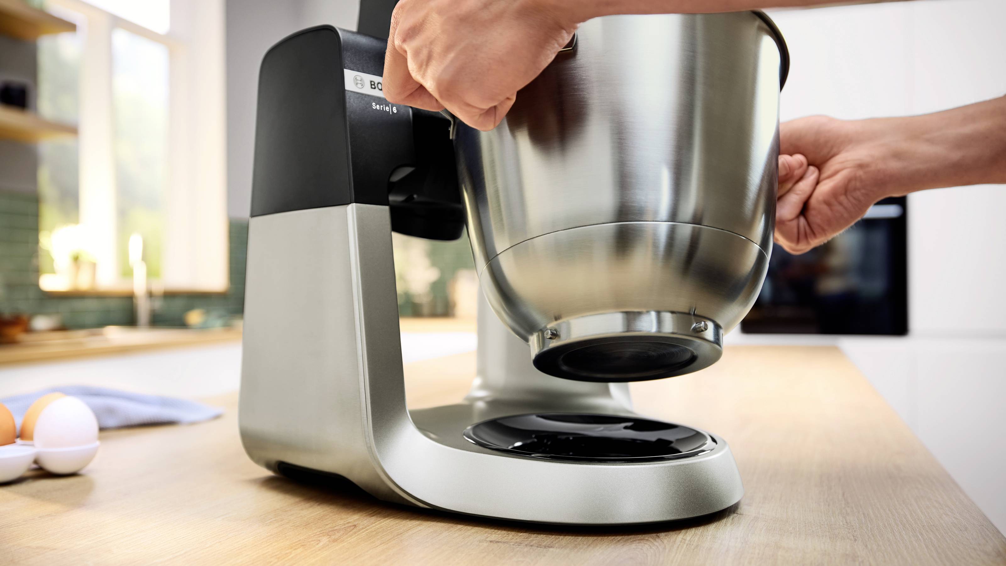 Een persoon die een roestvrijstalen mengkom op een moderne keukenmixer plaatst op een houten aanrecht. Eieren en een handdoek liggen in de buurt.