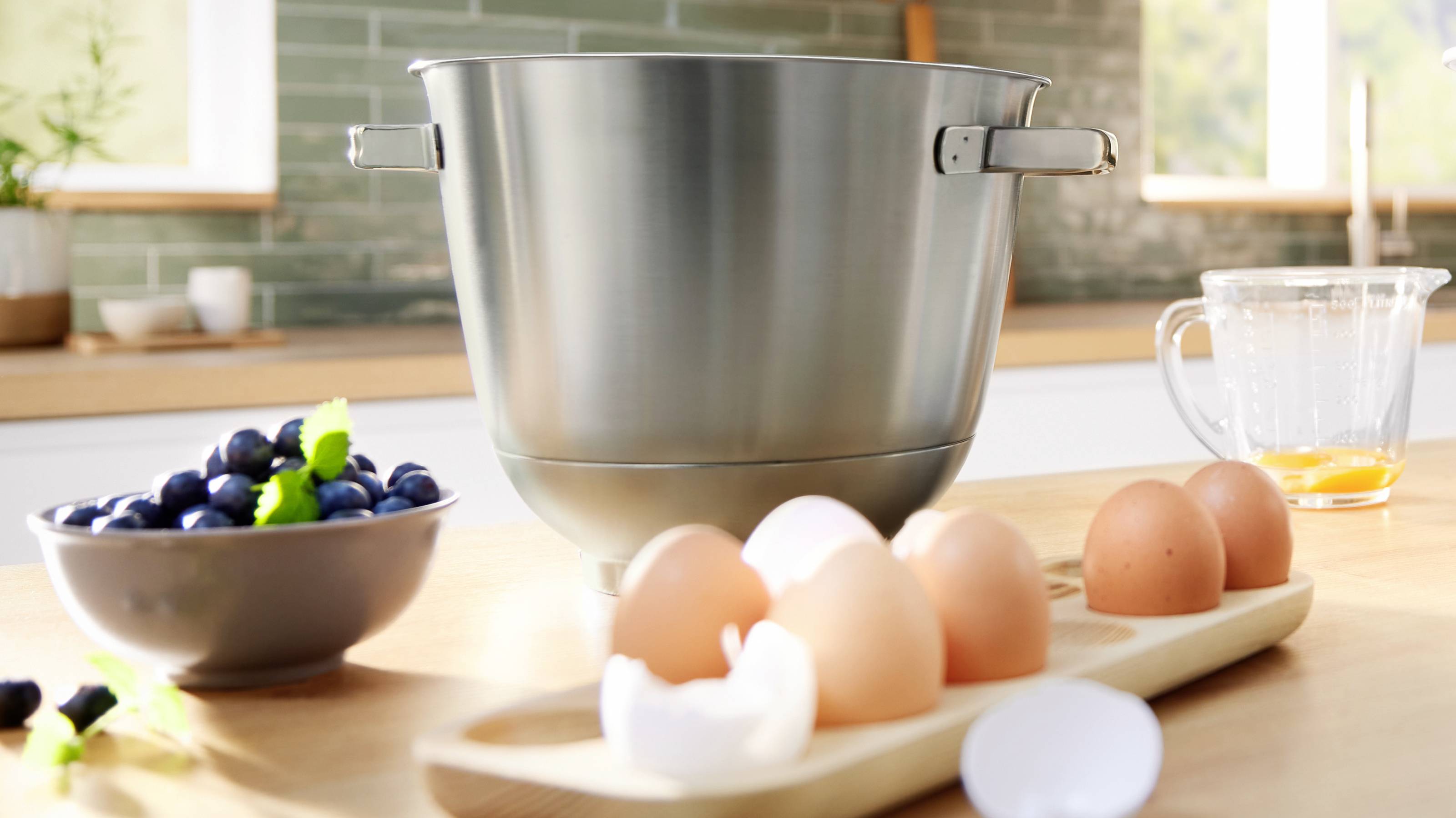 Een kom, eieren, bosbessen en een glazen kan met vloeistof op een aanrecht in een keuken met een raam op de achtergrond.