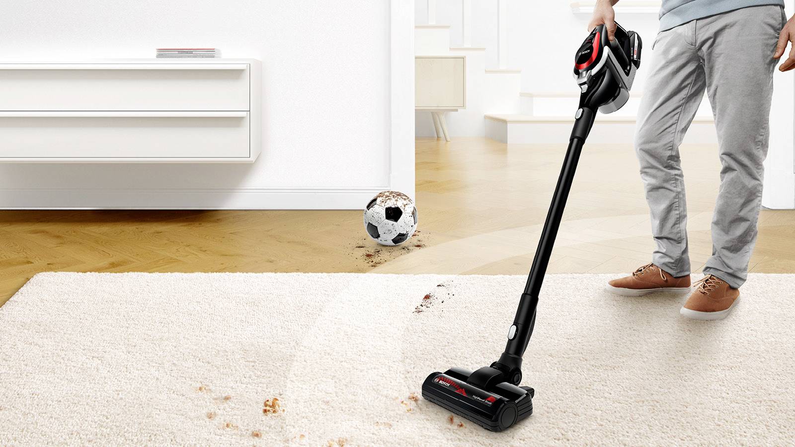 Een persoon gebruikt een steelstofzuiger op een beige vloerkleed en zuigt vuil op in de buurt van een voetbal.